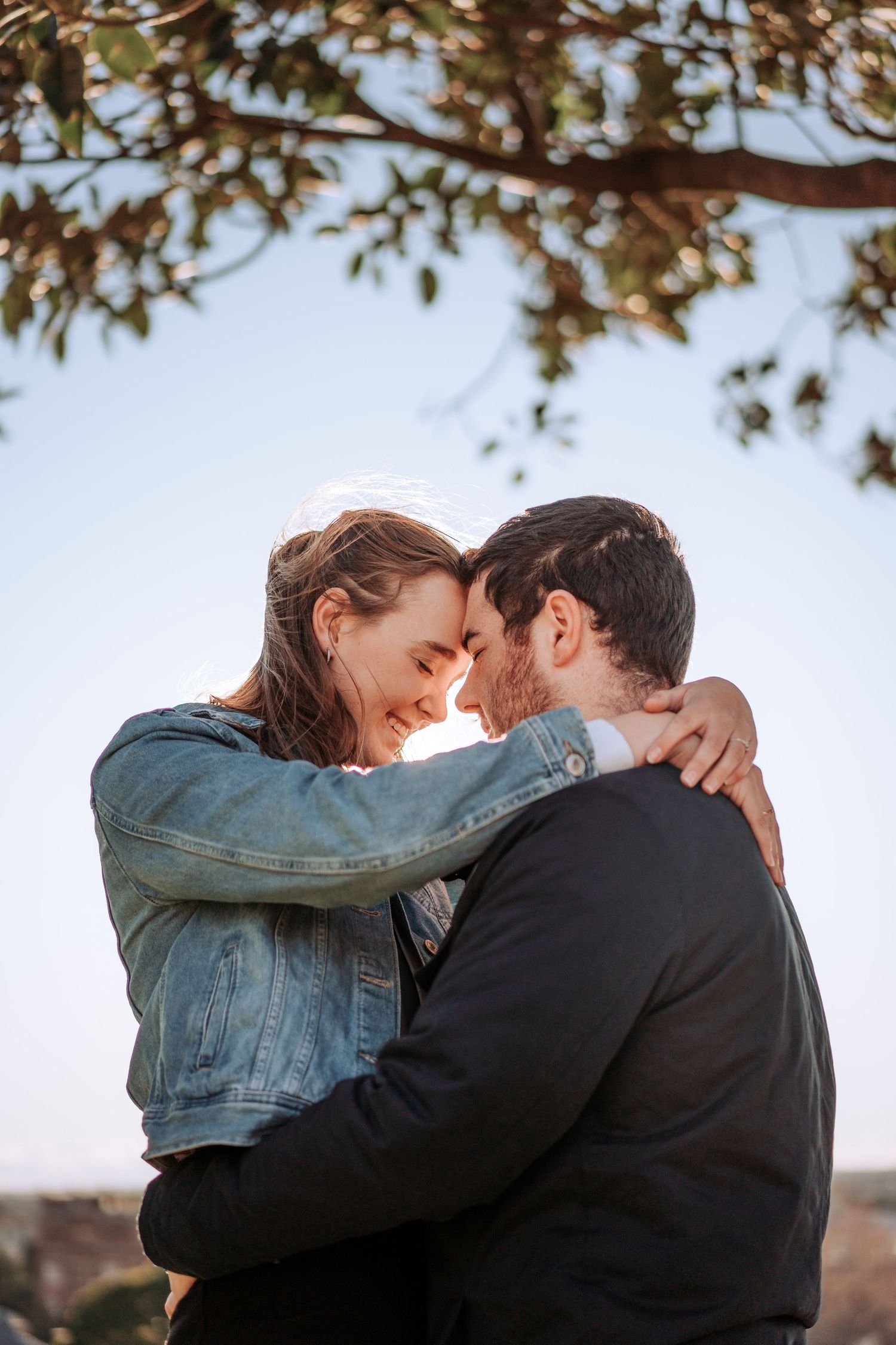 A romantic golden hour moment shows a couple embracing outdoors under tree branches against a bright sky.