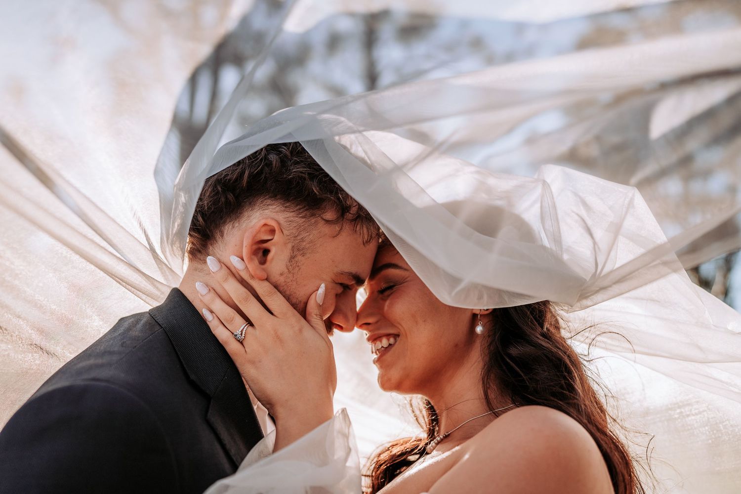 Romantic wedding couple embraces under flowing white veil during outdoor photo session.