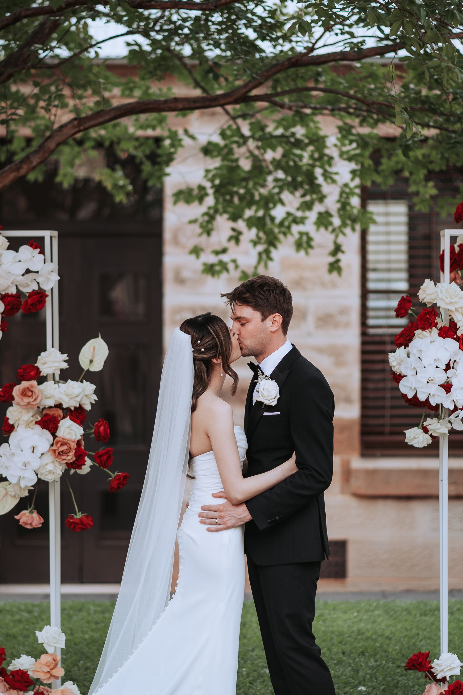A romantic garden wedding ceremony with floral arrangements featuring red and white flowers as a couple embraces.