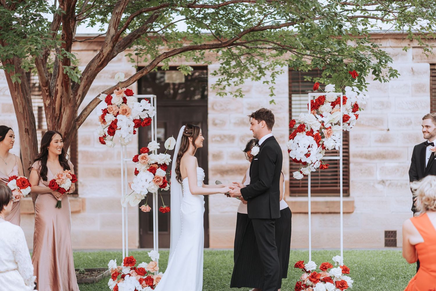 Outdoor wedding ceremony under a tree with red floral decorations and blush bridesmaids dresses.