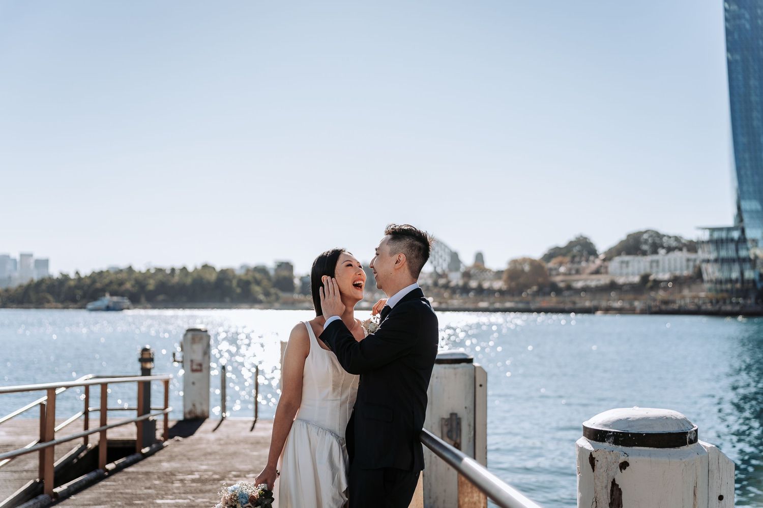 Couple sharing a romantic moment on a waterfront pier in wedding attire with city skyline and sparkling harbor behind them.