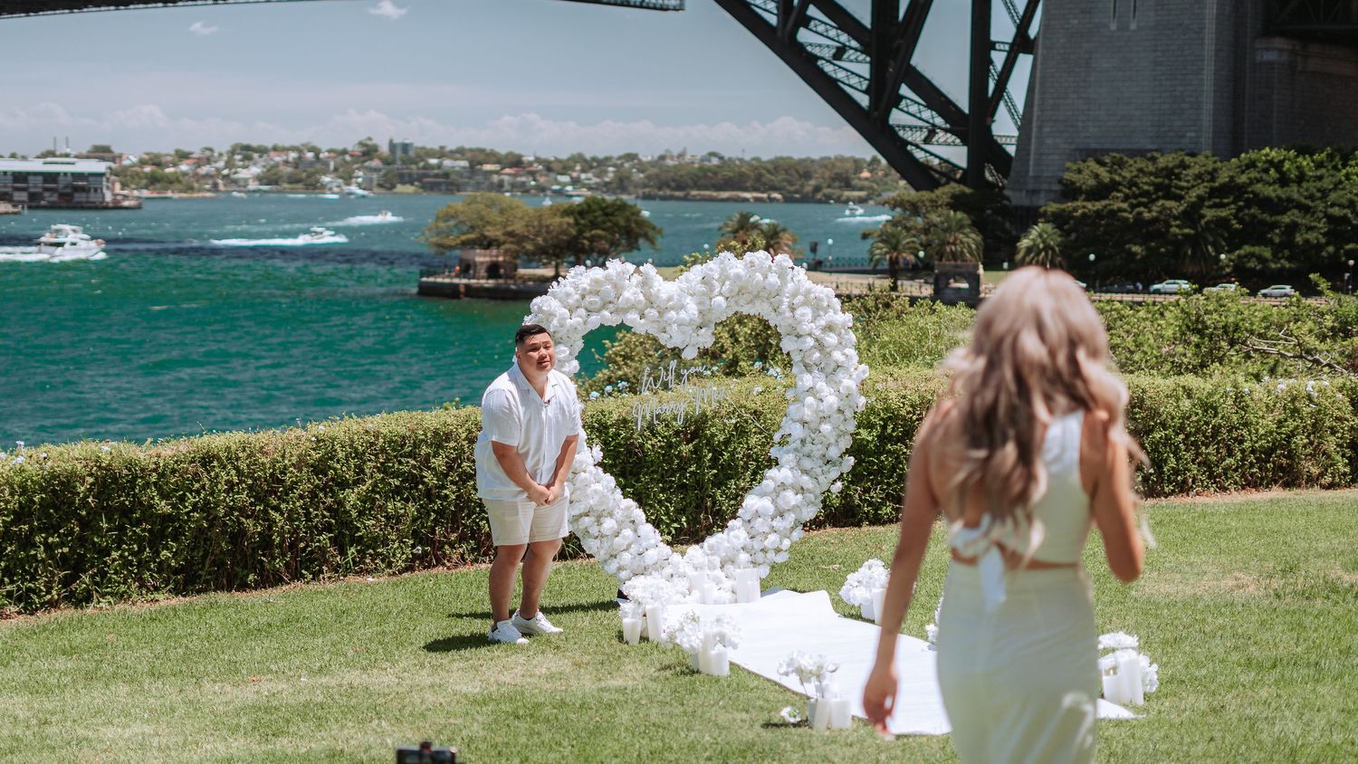 A couple creates a heart-shaped wedding photo on a grassy lawn overlooking turquoise waters in Sydney Harbour.