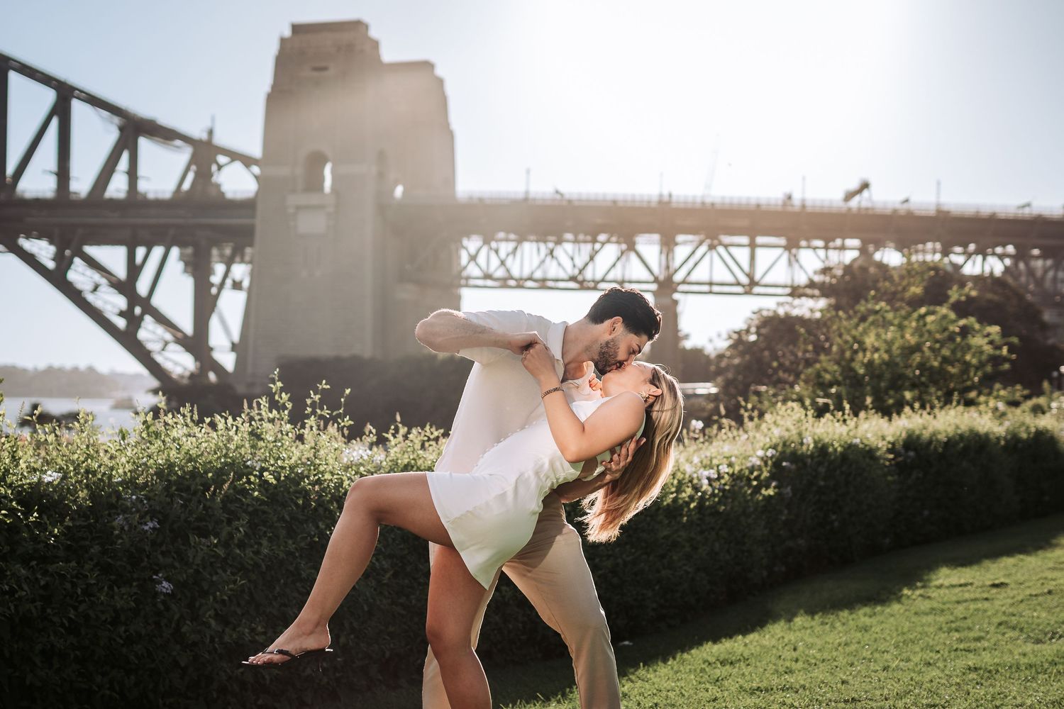 Romantic couple embrace in front of Sydney Harbour Bridge on a sunny afternoon with flowering bushes nearby.