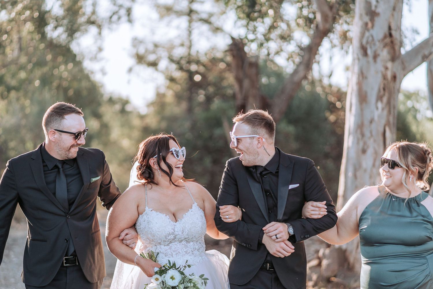 A joyful wedding party walks arm in arm outdoors under eucalyptus trees on a sunny afternoon.