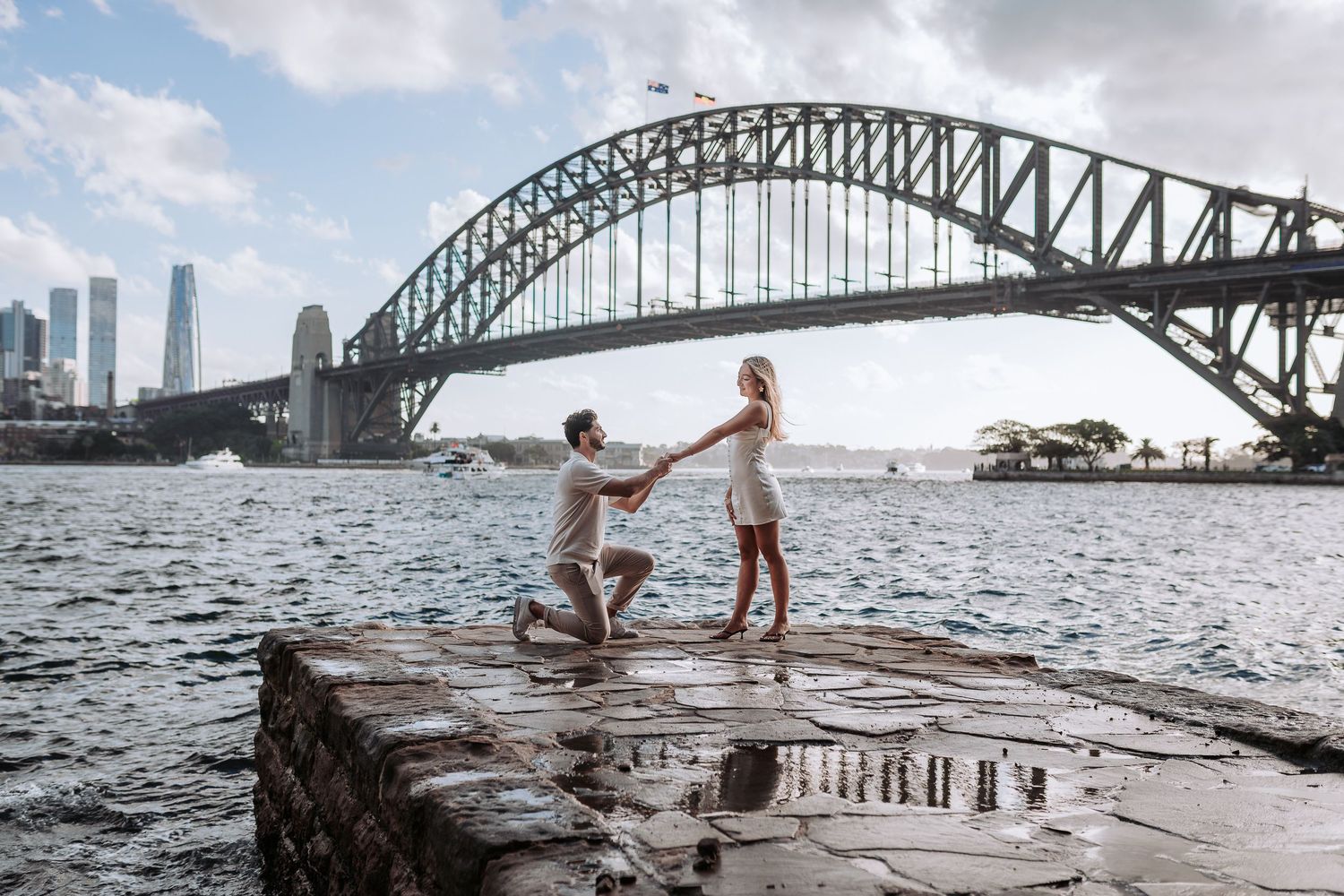 Man proposes to woman on rocky waterfront with Sydney Harbour Bridge backdrop on a cloudy day.