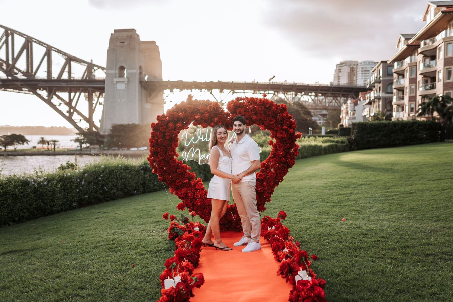 A couple stands in a heart-shaped red floral arch on an orange carpet with Sydney Harbour Bridge in the background.