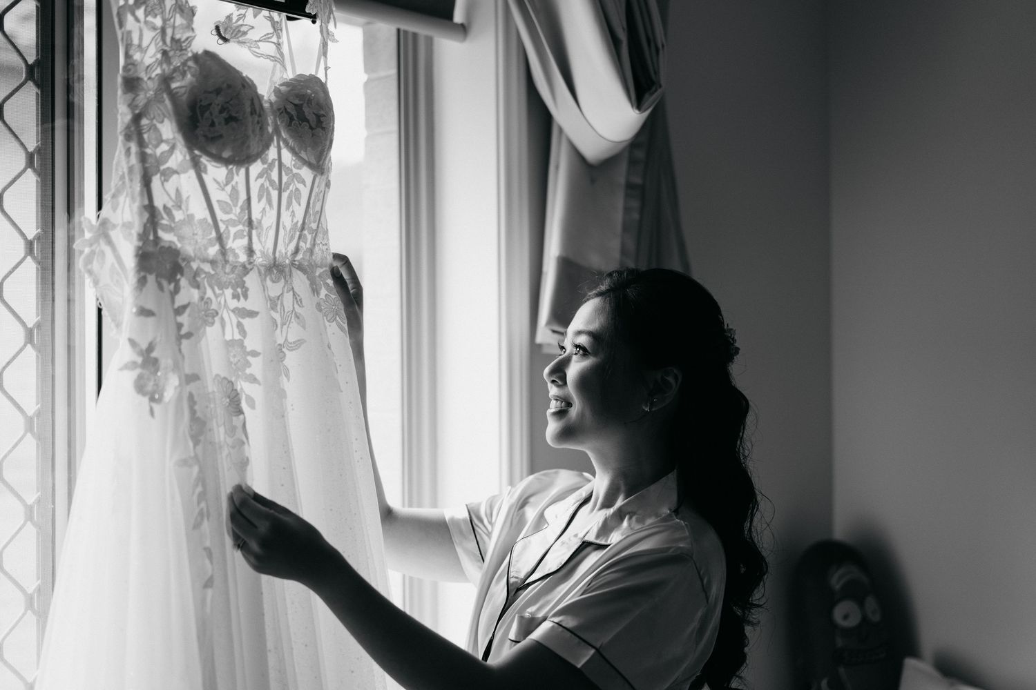 A wedding dress hangs by a window while a person in a robe admires it in black and white.