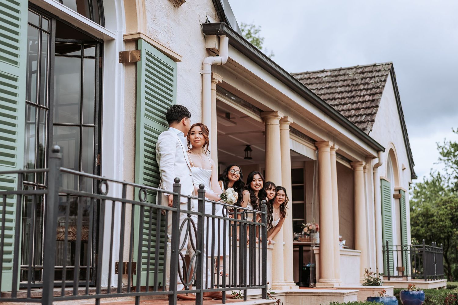 A group of people pose together on a balcony of a traditional Louisiana-style home with green shutters and columns.