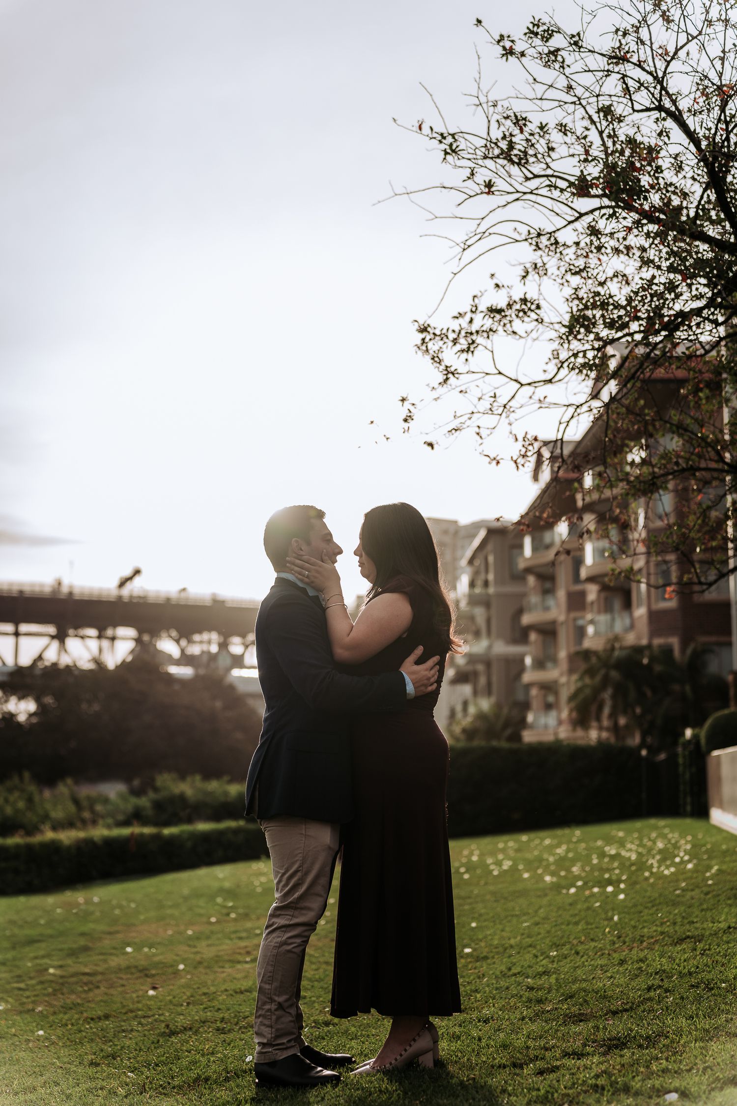 Couple embracing at golden hour in a lush park with a building and bare tree branches in the background.