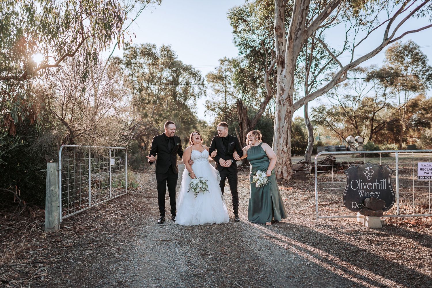 Wedding party walks down a sunlit dirt path between eucalyptus trees with a welcome sign visible at outdoor venue.