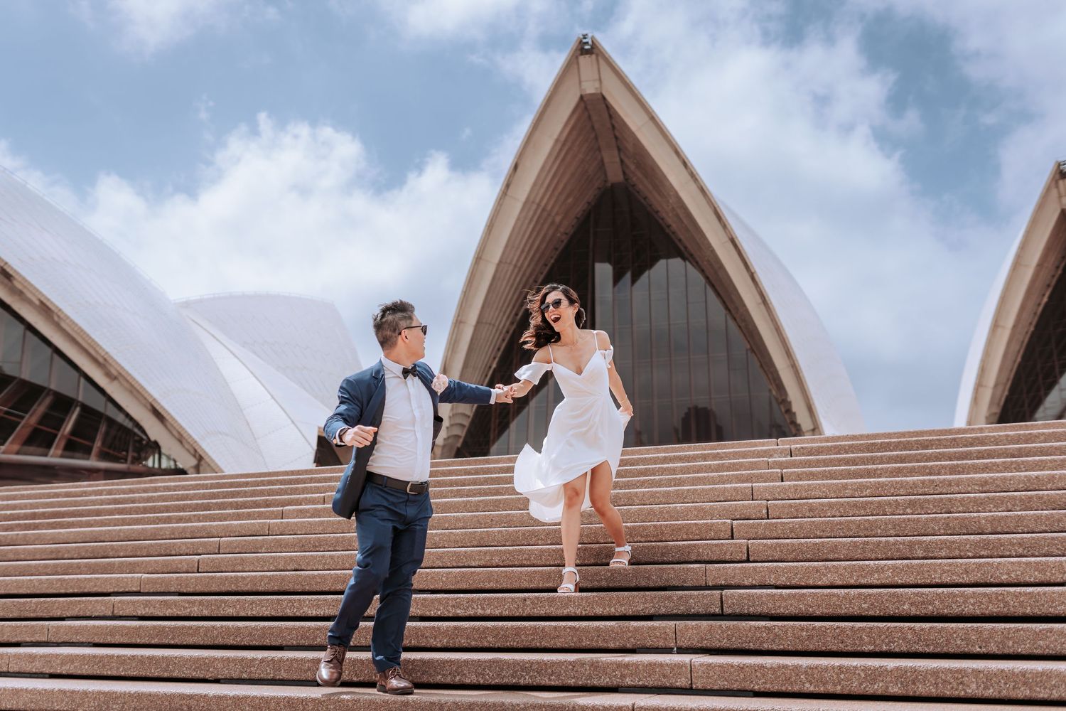 A couple holds hands on the steps of the iconic Sydney Opera House on a sunny afternoon in Australia.