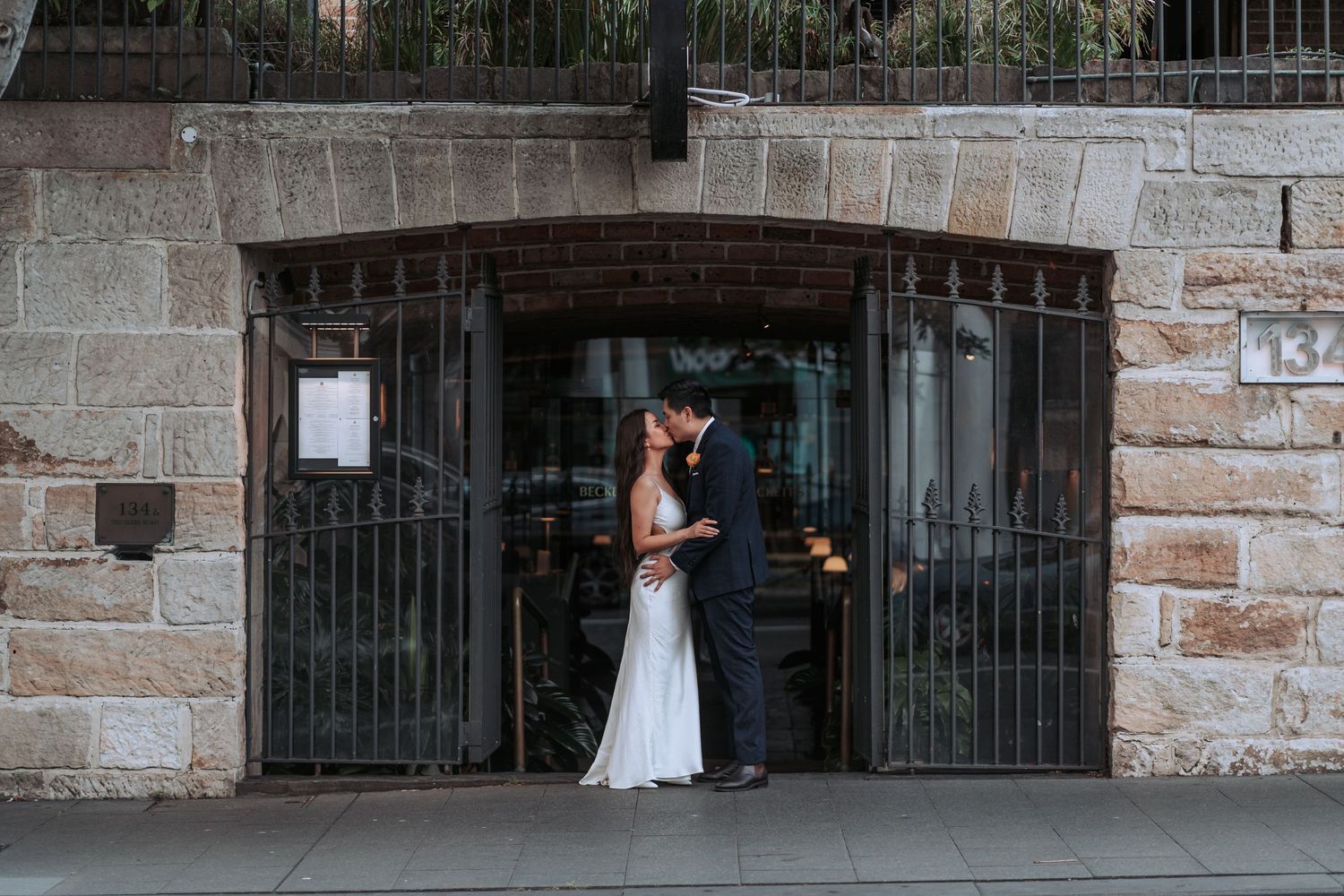 A couple in wedding attire embraces in front of rustic stone archway with ornate metal gates.