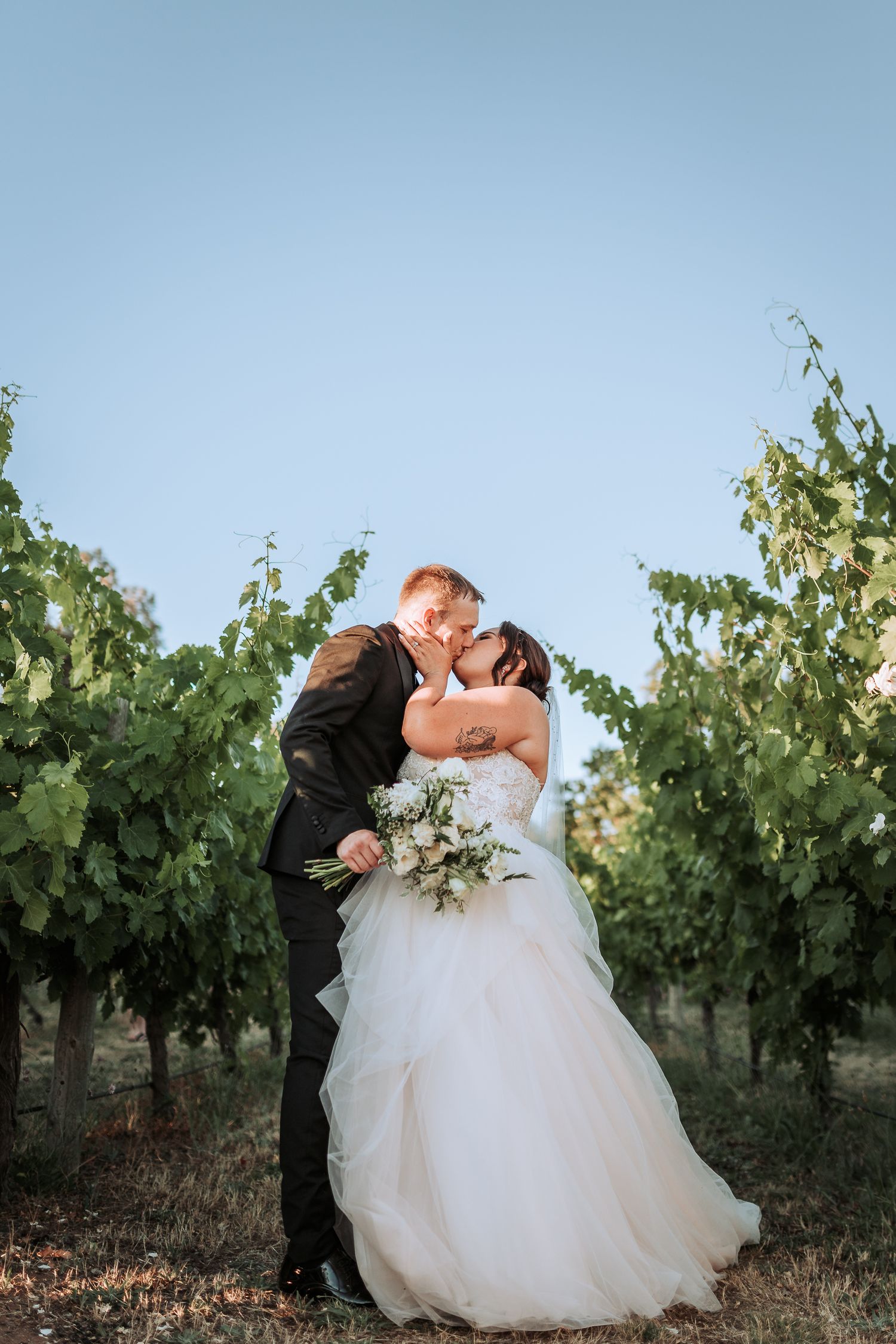 A romantic wedding couple embraces in a scenic vineyard at sunset as her white gown and bouquet glow in the soft light.