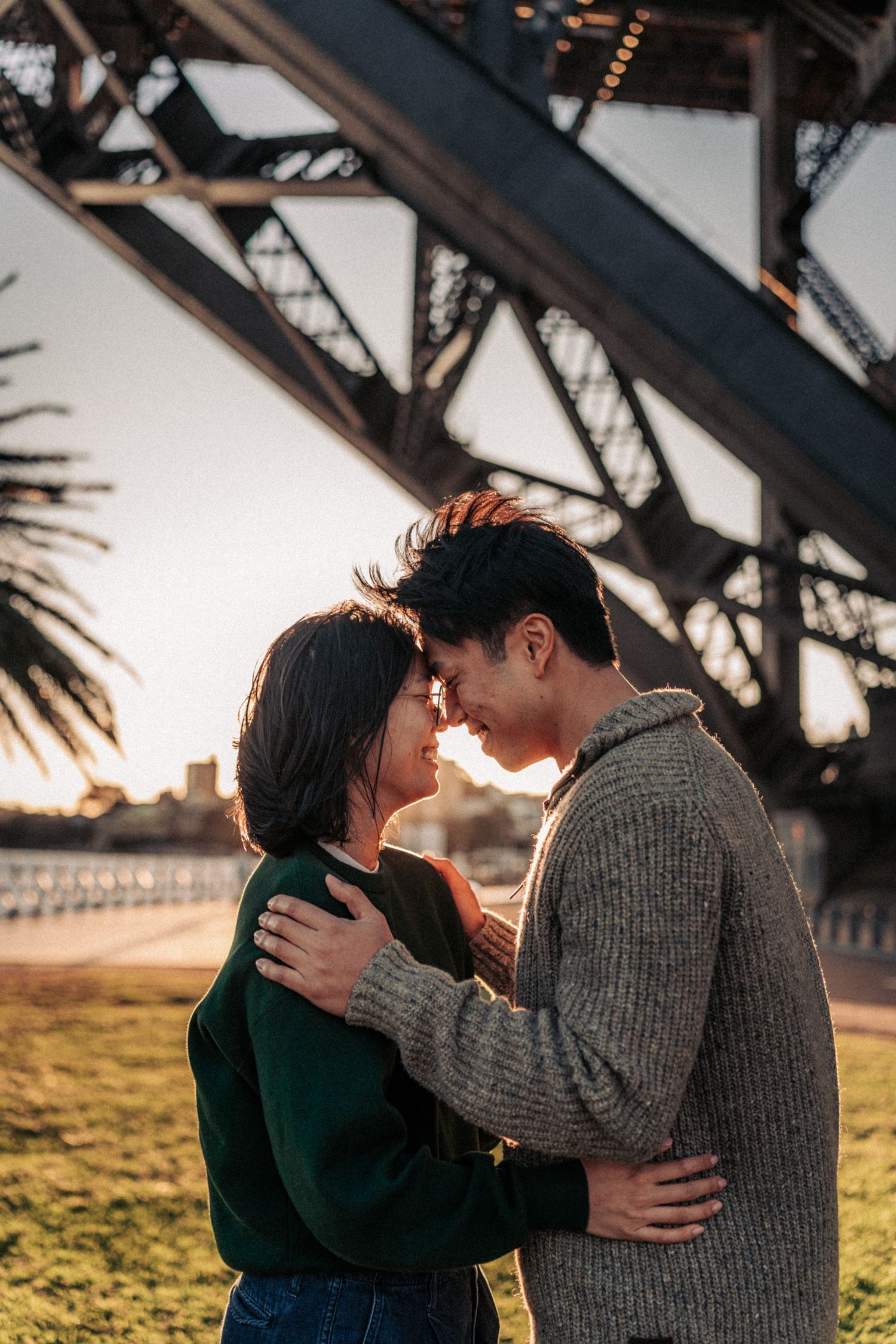 Couple embracing at sunset beneath Sydney Harbour Bridge, sharing a romantic moment on the grassy waterfront.
