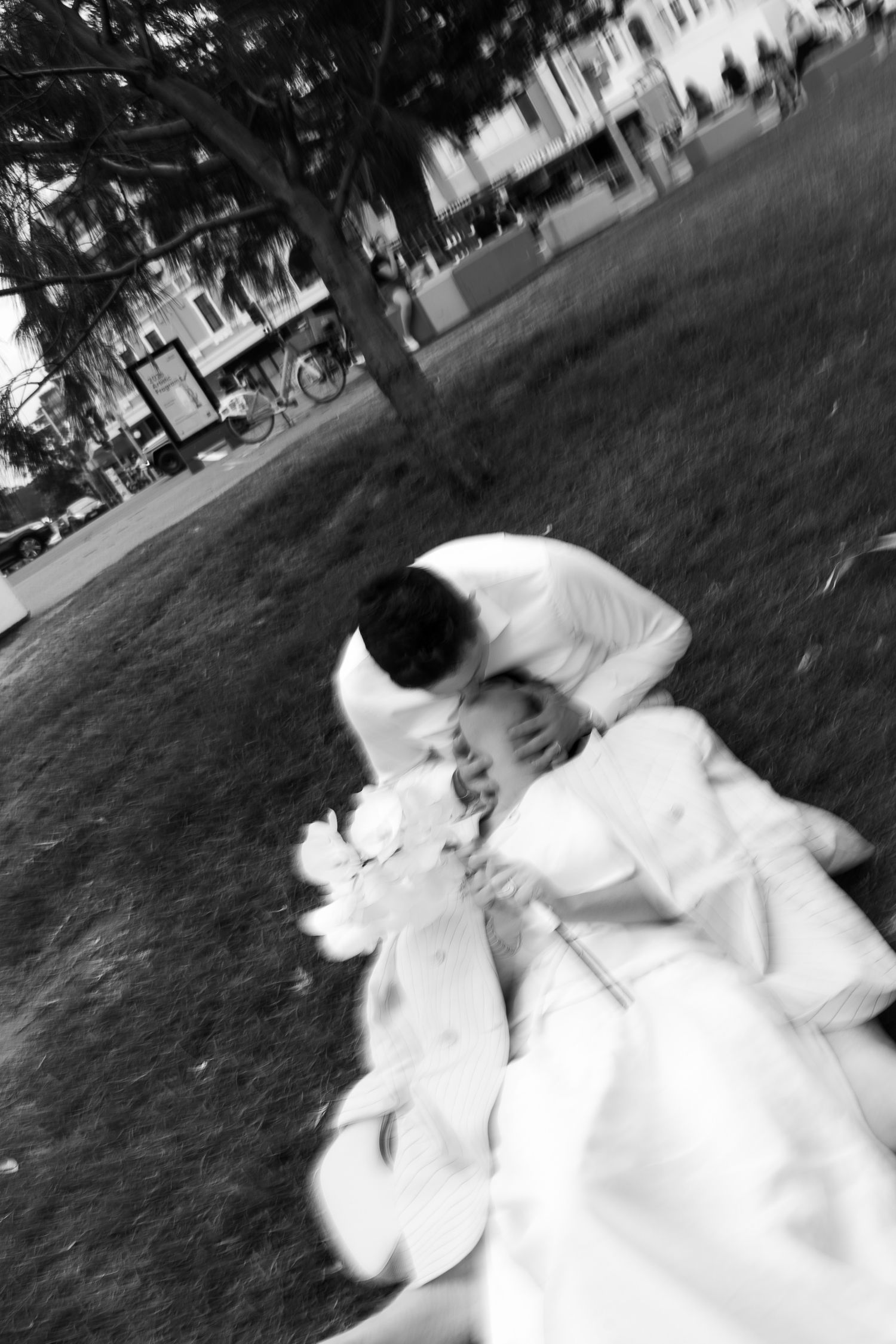 Black and white photo of a bride and groom sharing a romantic moment on grass, with trees and buildings in the background.