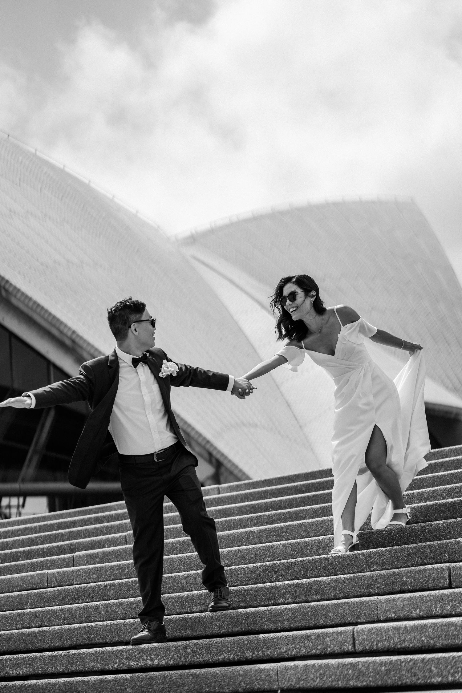A couple in formal attire runs playfully down the steps of the Sydney Opera House in black and white photography.