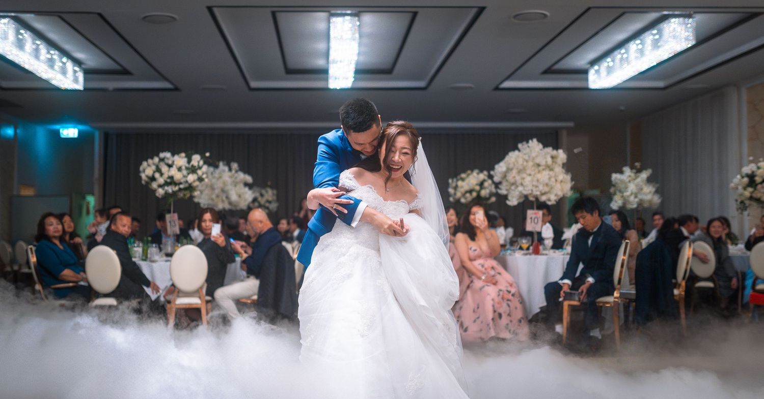 A couple sharing their first dance at a wedding reception with romantic fog effects on the dance floor and white floral decor.