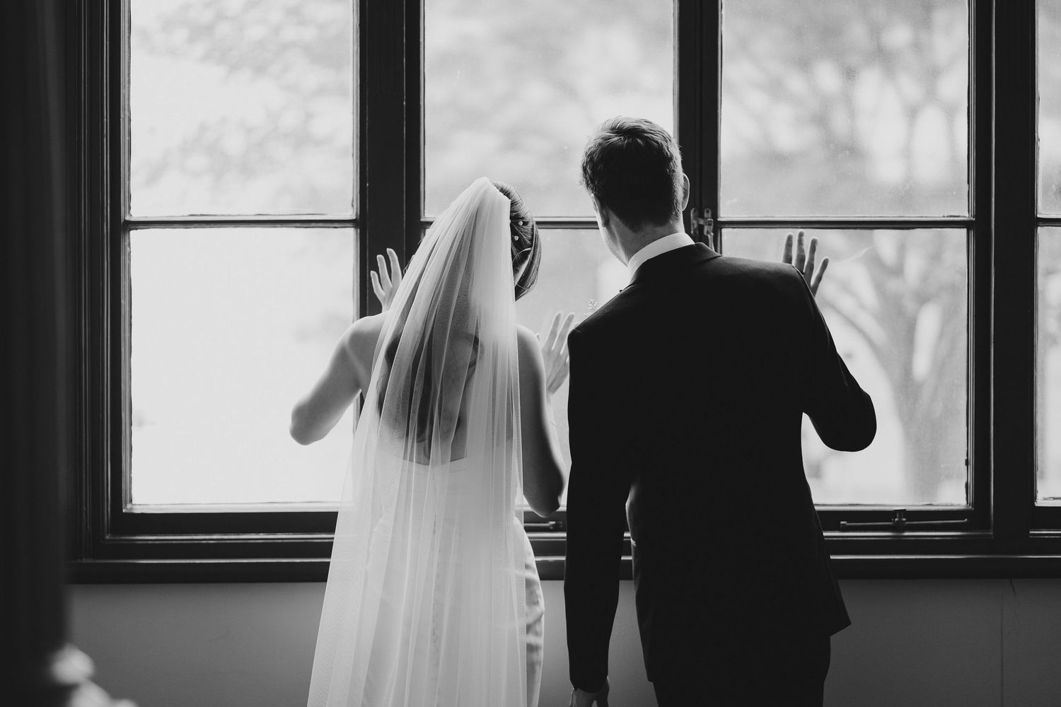 A black and white silhouette of a couple in wedding attire looking out a large window together.