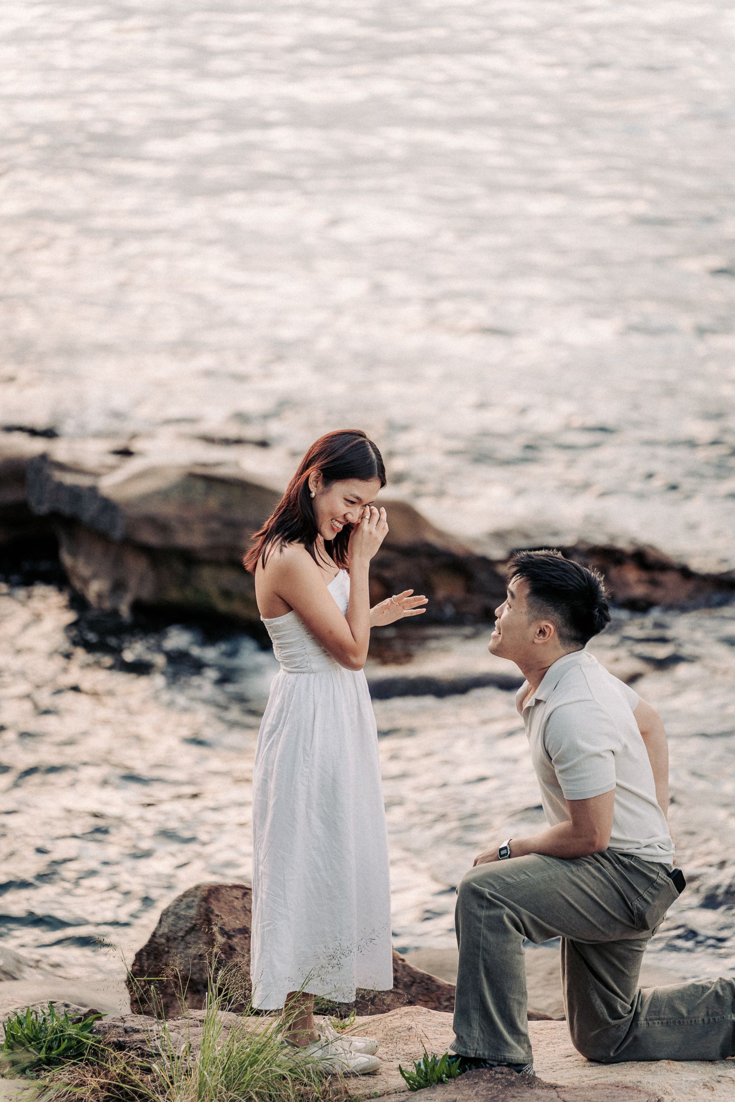 Man proposes on one knee to emotional woman in white dress by rocky seaside at sunset.
