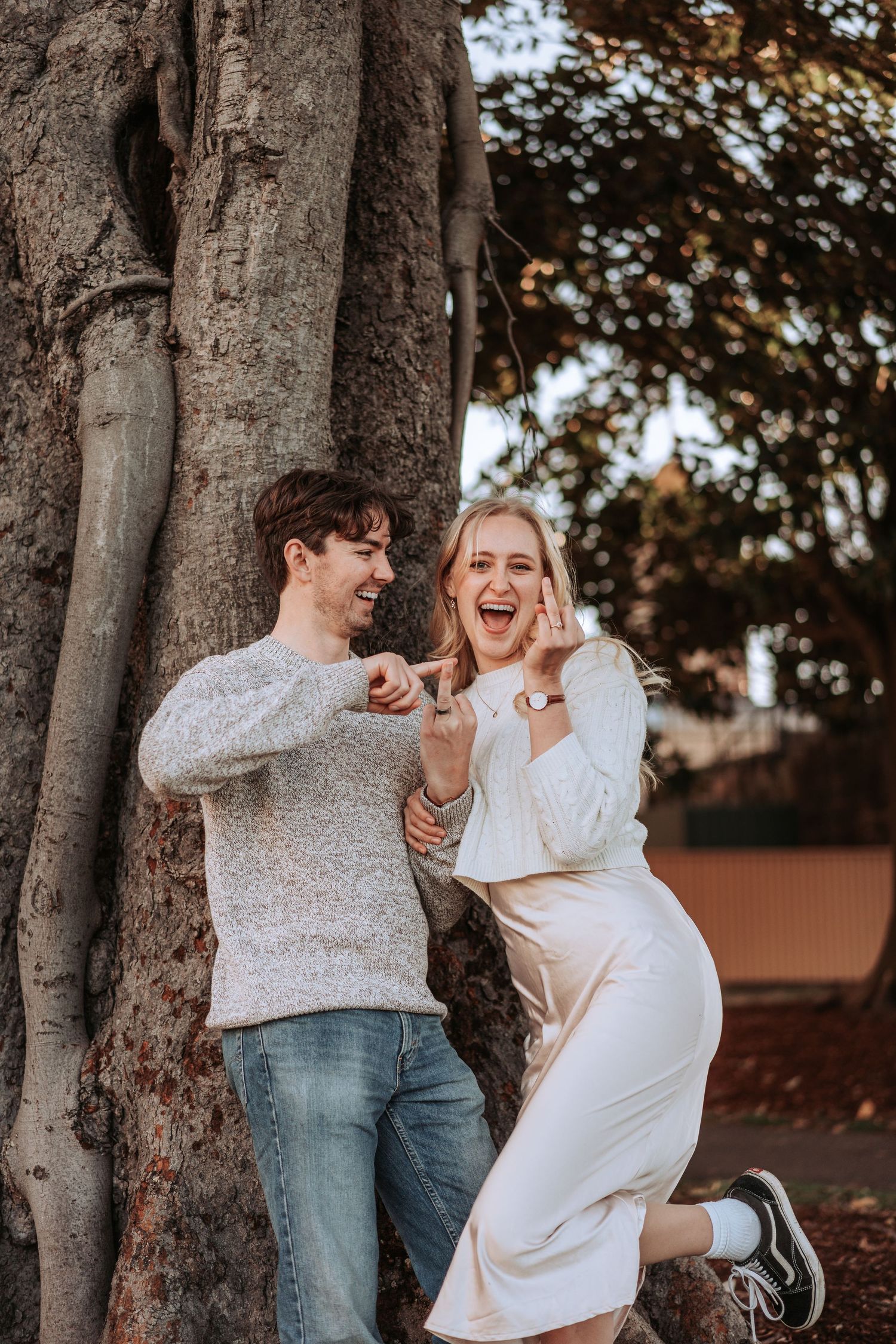 A couple in white sweaters laughs joyfully while leaning against a large tree trunk in autumn lighting.