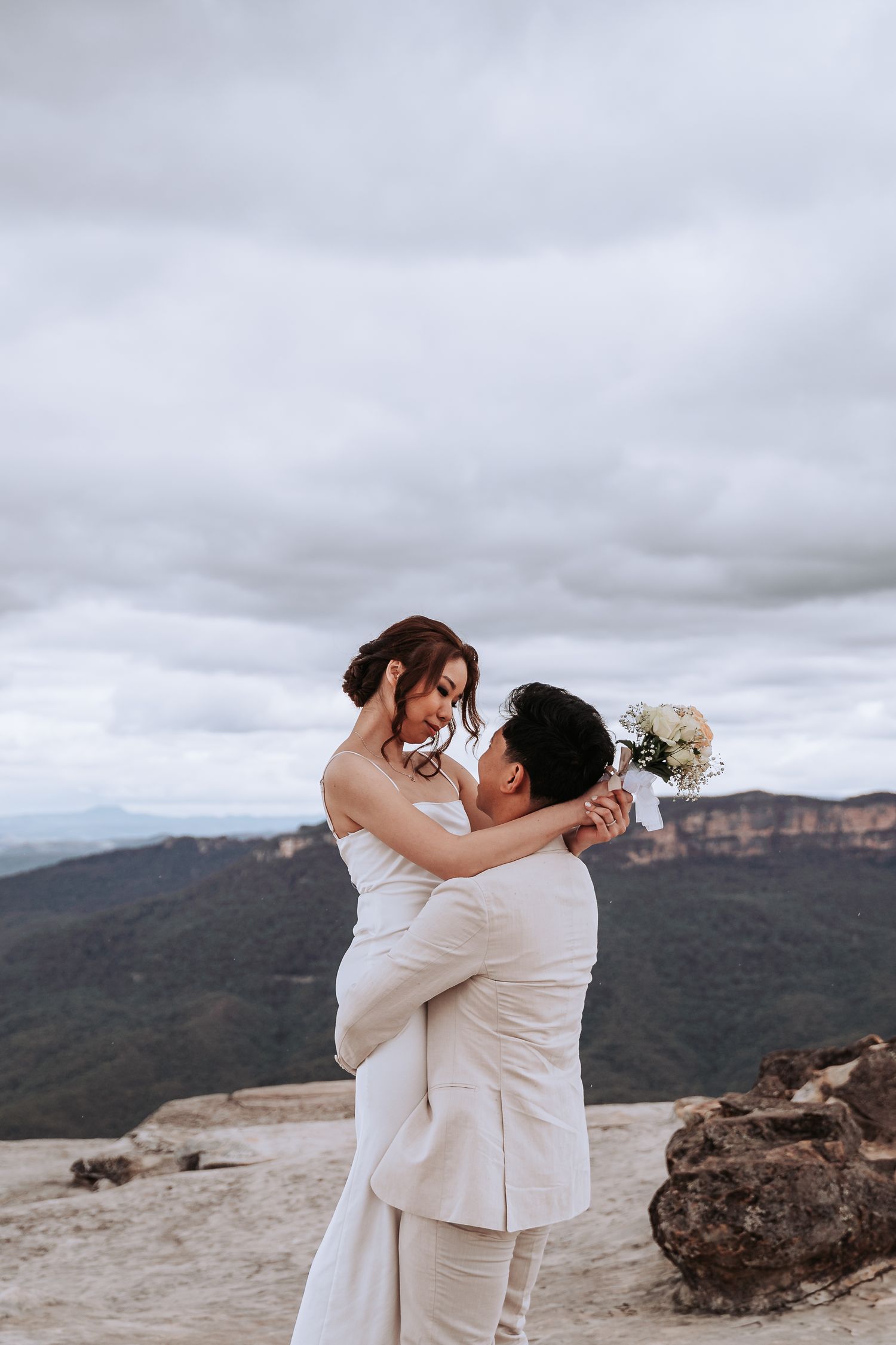 A romantic wedding couple embracing on a mountaintop with cloudy skies and mountains in the background.