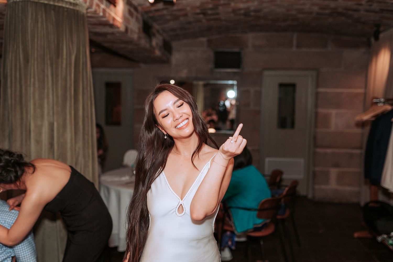 A person in a white dress dancing and smiling at a reception venue with dim lighting.