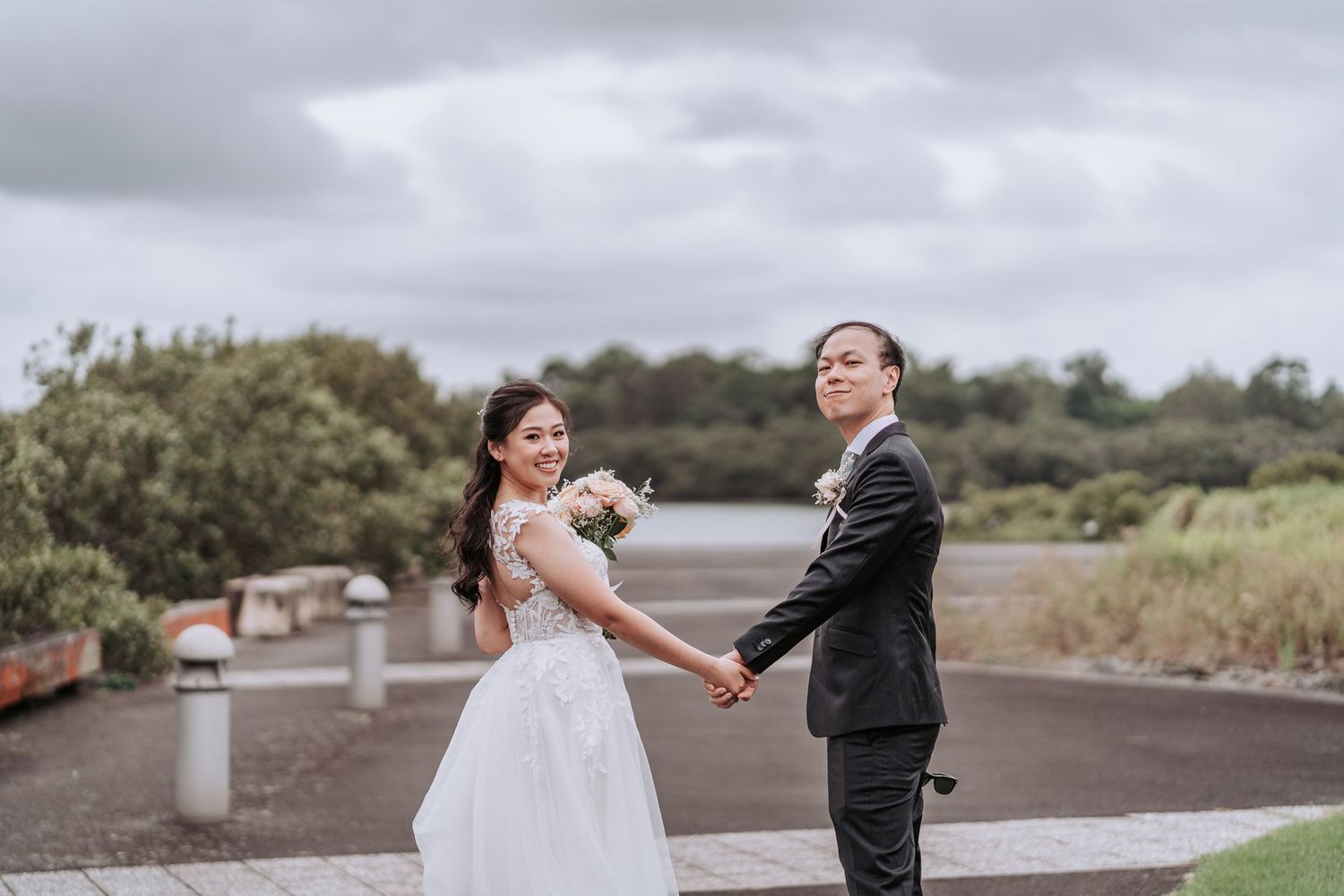 A couple in wedding attire holds hands and smiles on a winding path under cloudy skies.