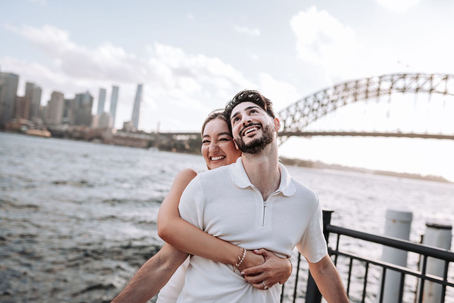 A couple shares a joyful embrace while standing by the harbor with Sydney Harbour Bridge in the background.