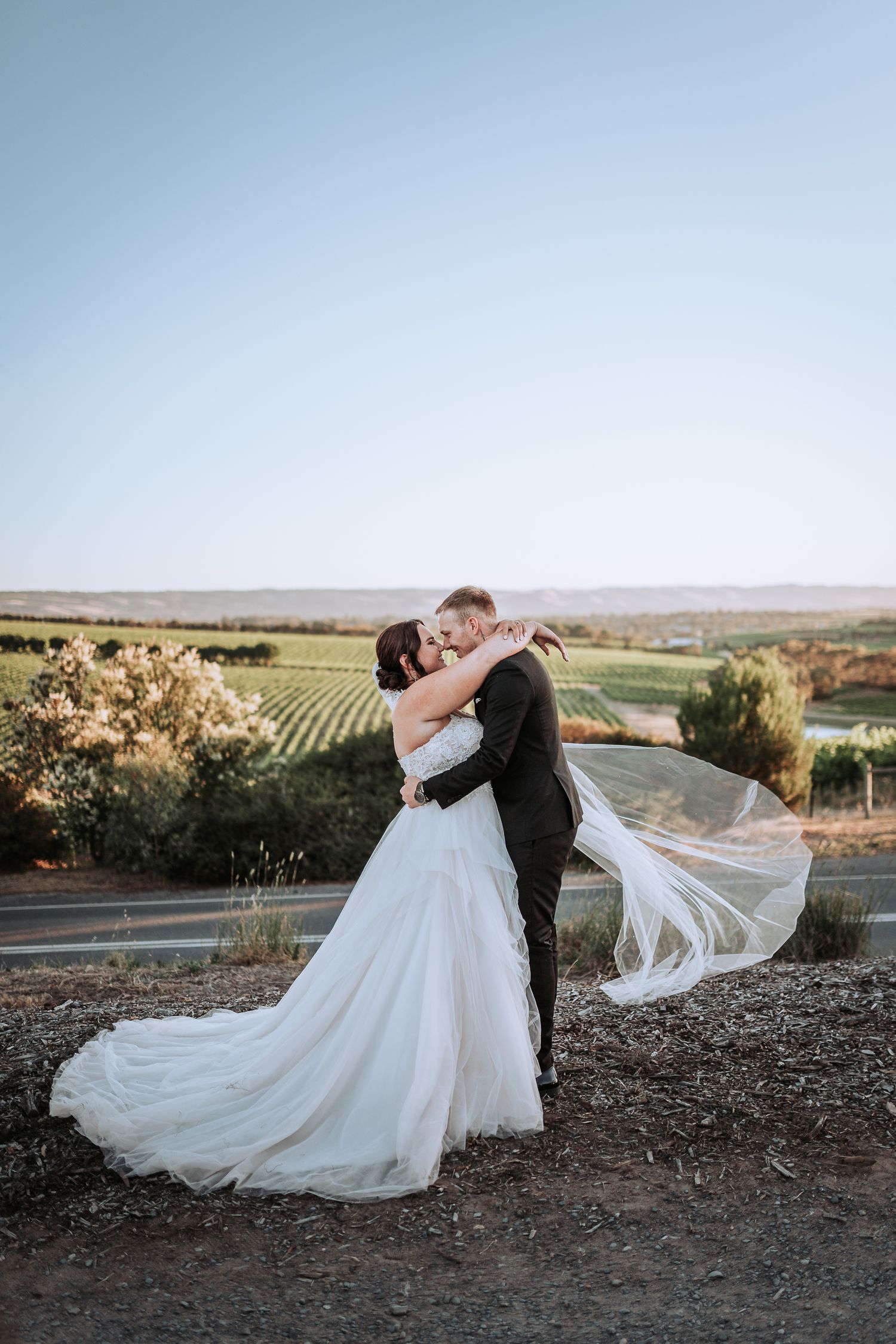 A couple shares an intimate moment at sunset overlooking vineyard rows in a scenic winery setting.