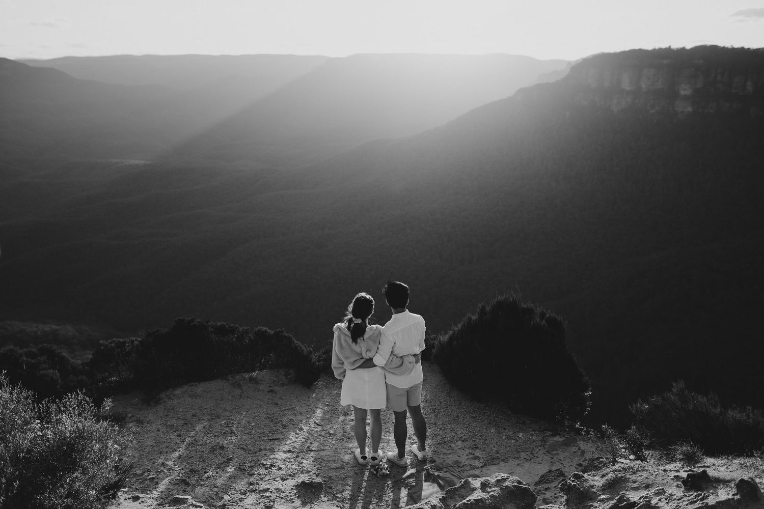 A couple stands silhouetted against a dramatic mountain vista during sunset in black and white photography.