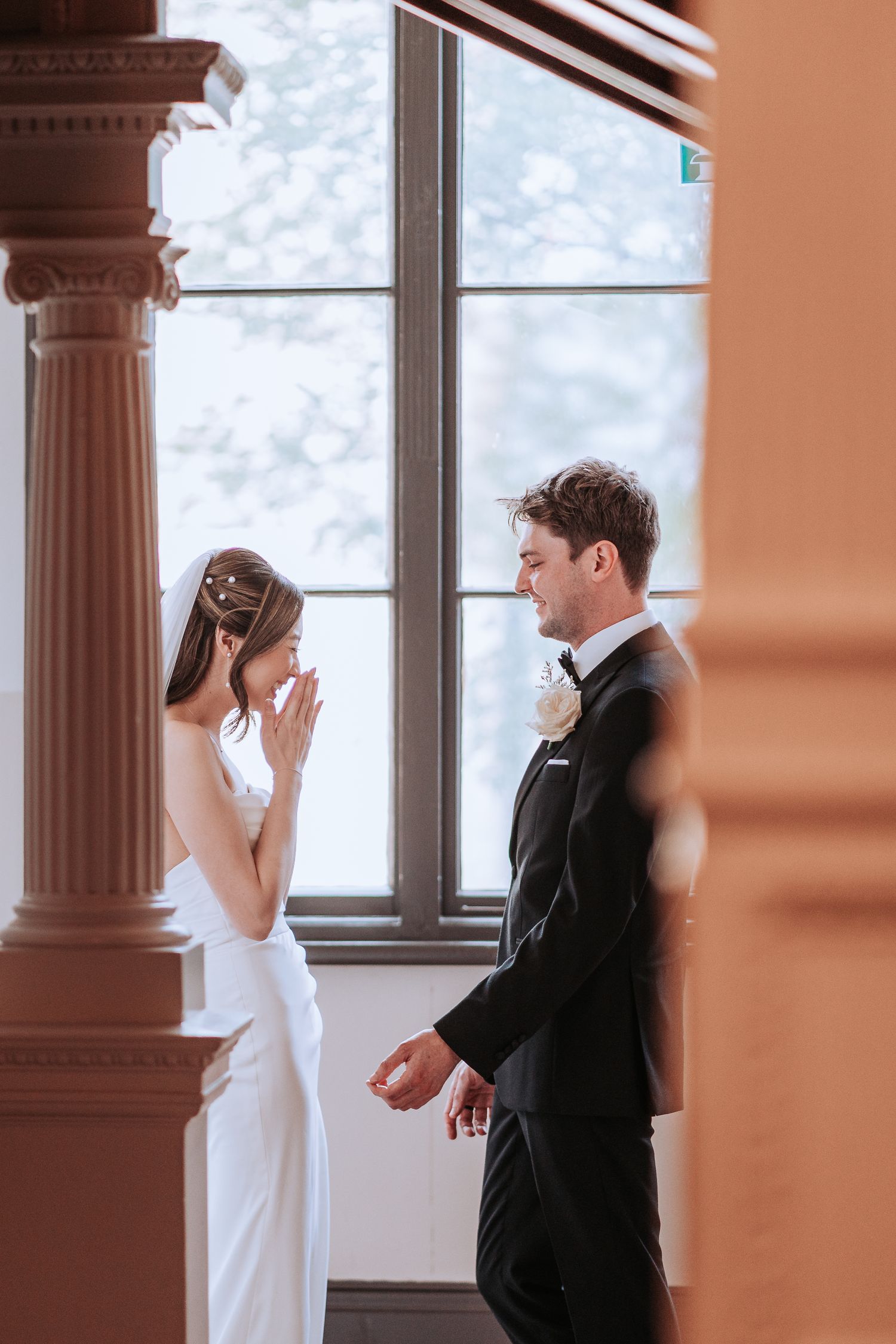 A private first look moment between newlyweds in formal attire by an elegant window with decorative pillars.