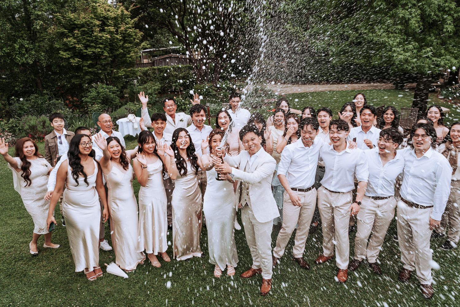 Wedding party in all white attire celebrating with champagne spray in an outdoor garden setting.