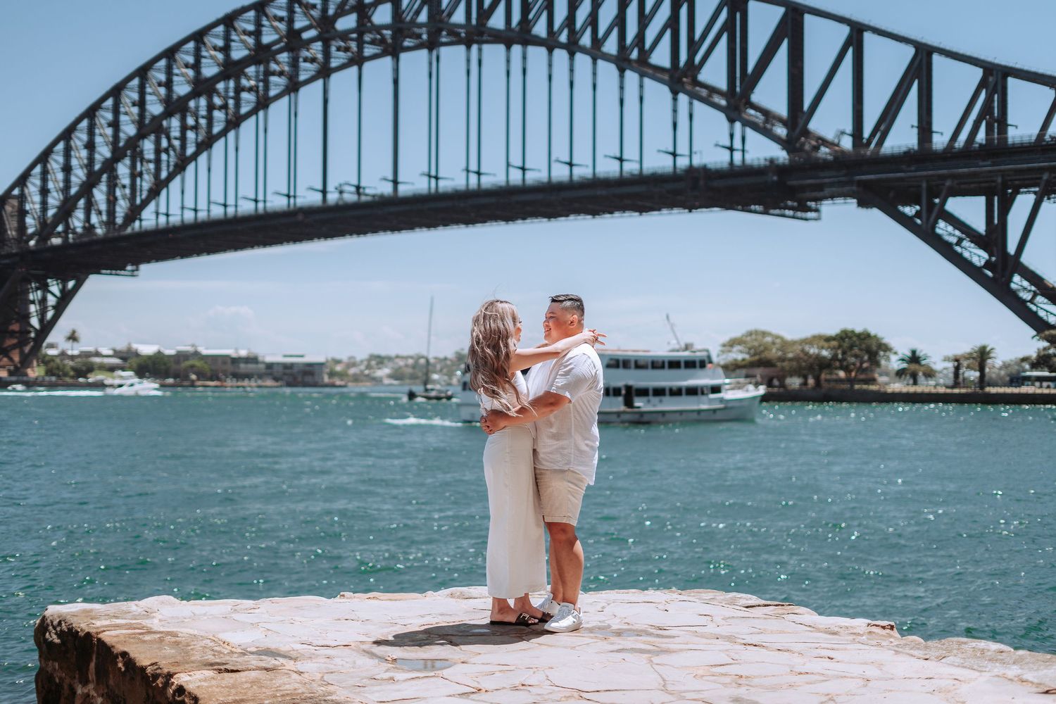 Two people embrace on a waterfront platform with the Sydney Harbour Bridge framing the romantic scene against blue water.