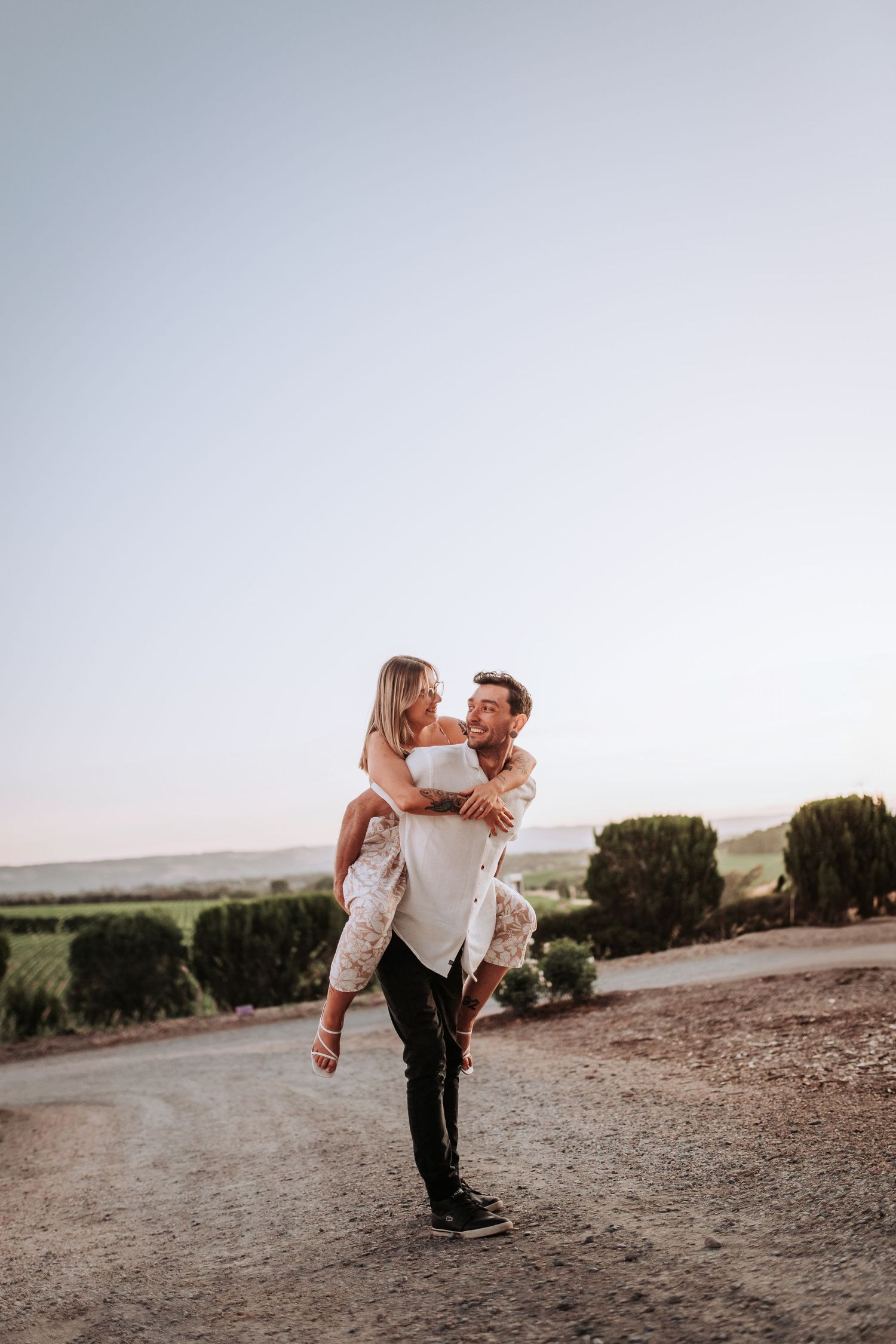 Couple in casual attire embracing during sunset with scenic rural backdrop.