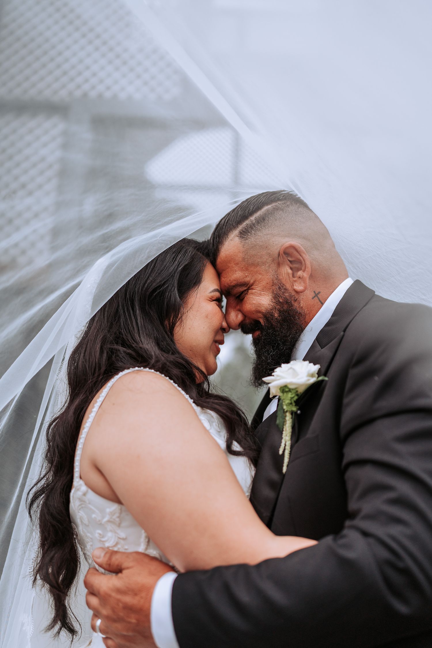 A happy newlywed couple shares an intimate forehead touch moment under a flowing white veil at their wedding.