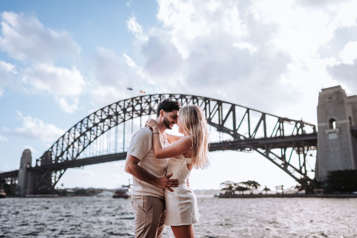 A romantic couple shares an intimate moment in front of the iconic Sydney Harbour Bridge on a sunny day.
