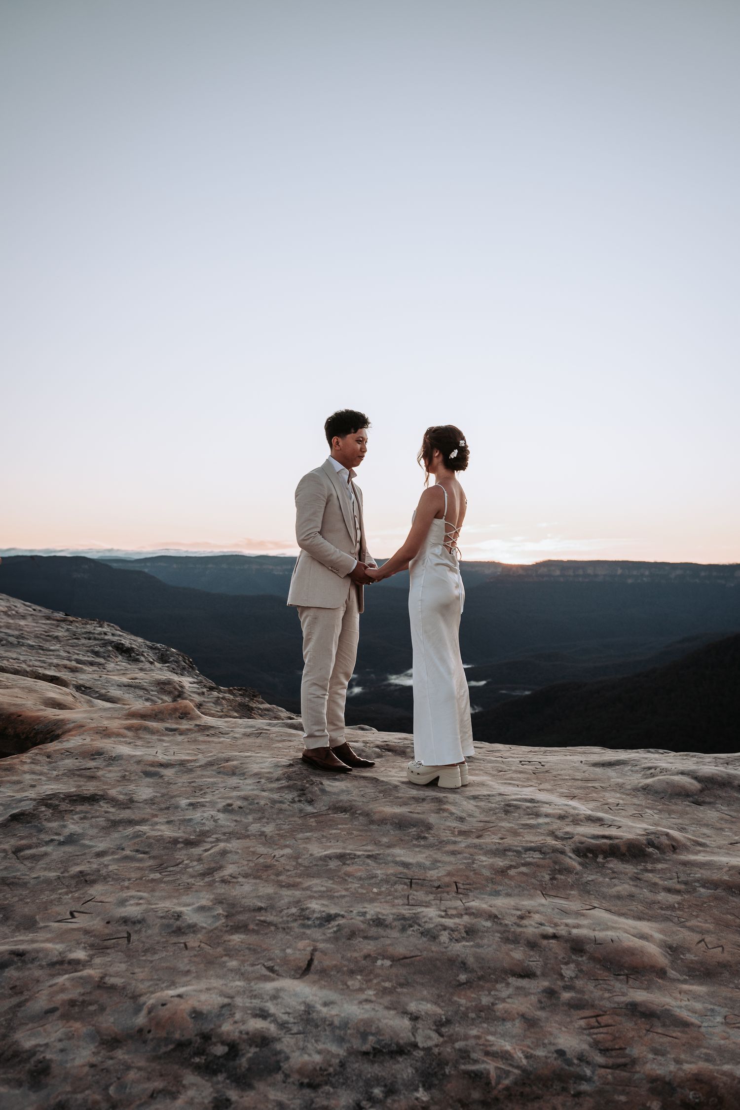 A couple in wedding attire stands on a rocky mountain overlook at sunset.