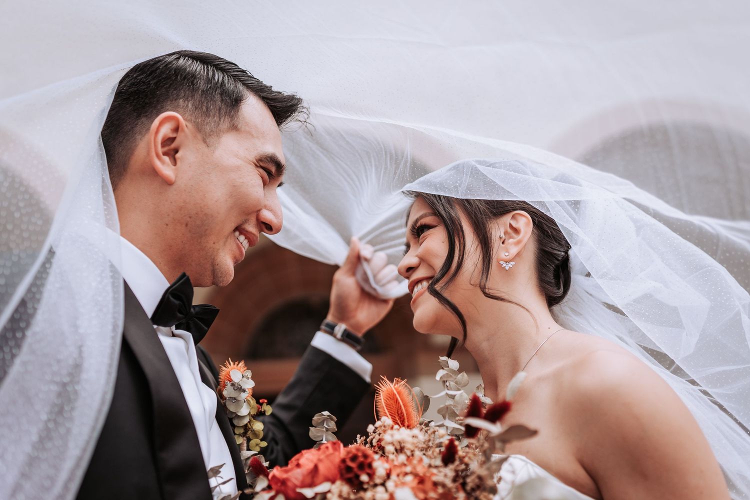 Close-up tender moment of newlyweds under veil with coral pink bouquet at wedding ceremony.