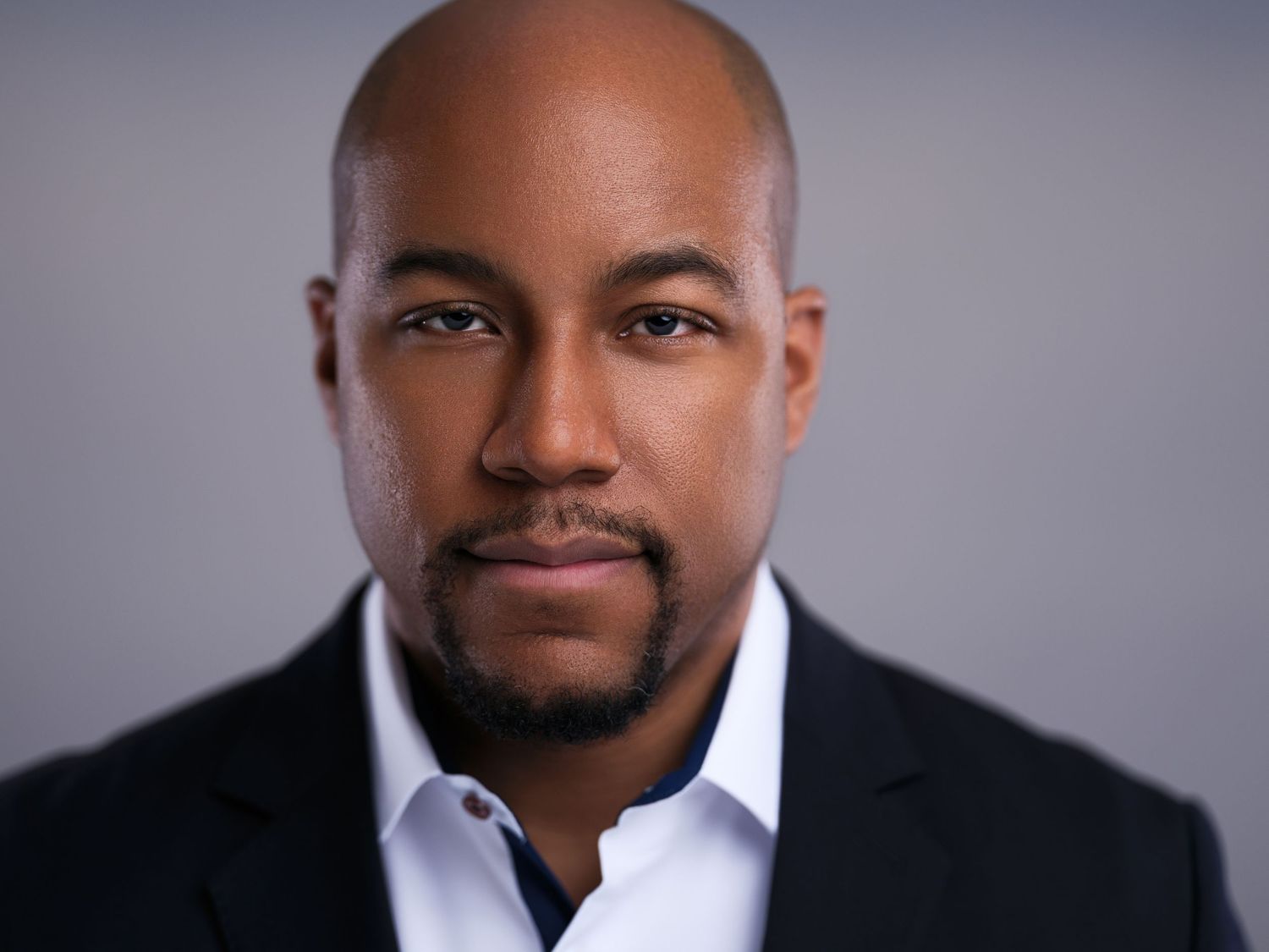 Professional headshot of a bald man in a black suit and white shirt against a grey backdrop.