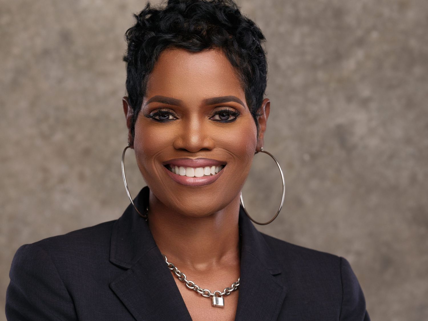 Professional studio portrait of someone wearing a black blazer, silver hoop earrings and necklace, with short dark hair and a bright smile.