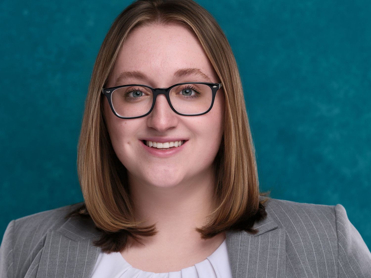 Professional headshot of someone wearing glasses and a grey pinstriped blazer against a teal background.