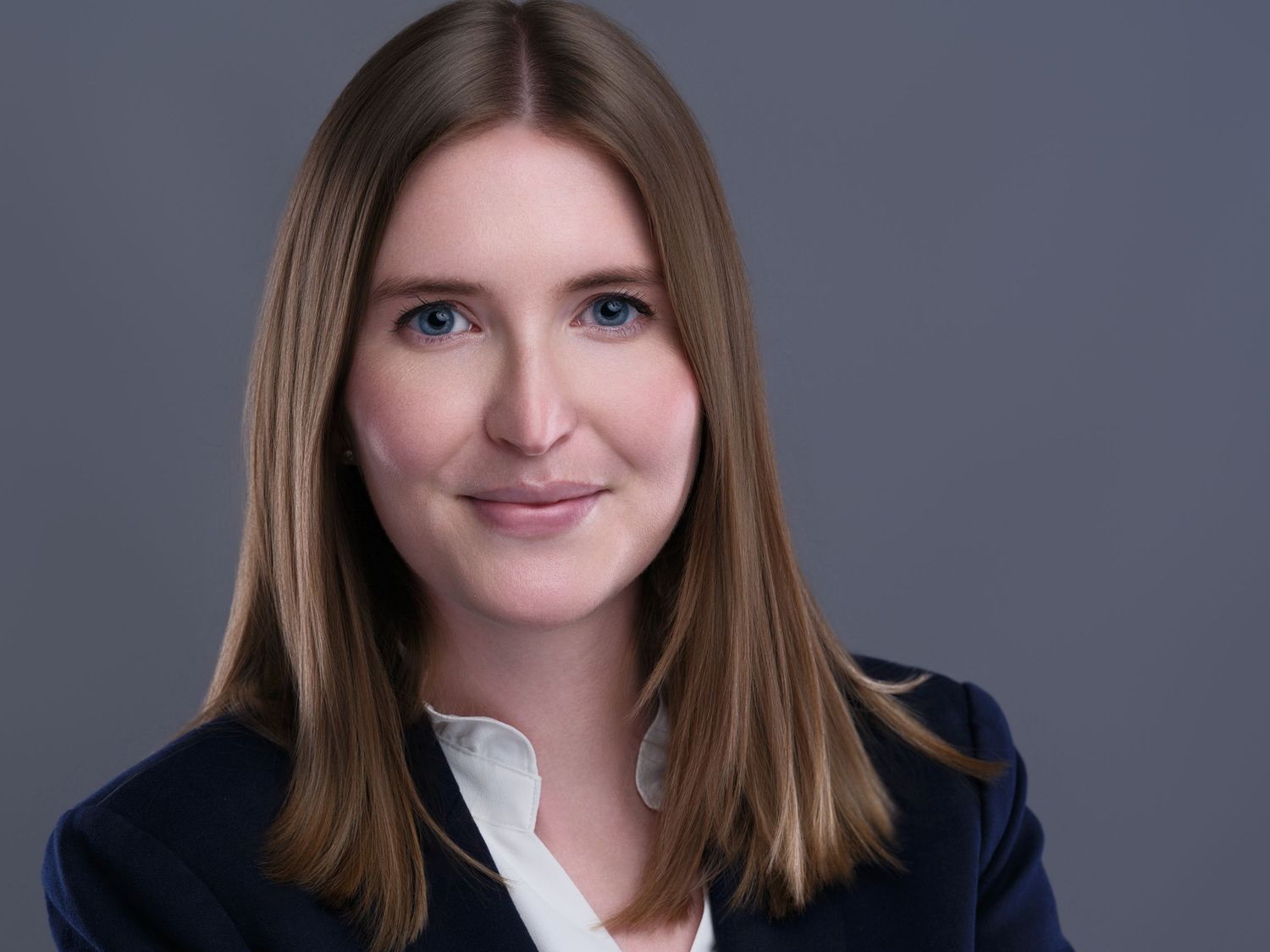 Professional headshot of a person with straight brown hair and a navy blazer against a gray background.