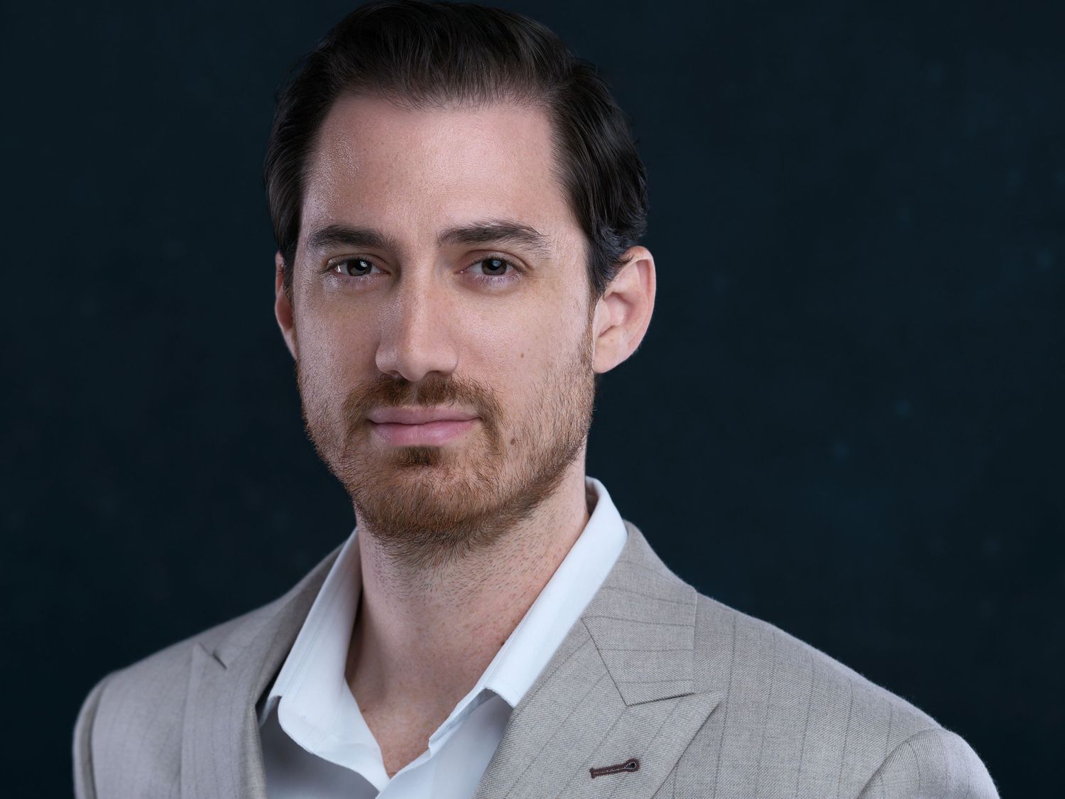 Professional headshot portrait against dark background showing person wearing light gray blazer and white shirt.