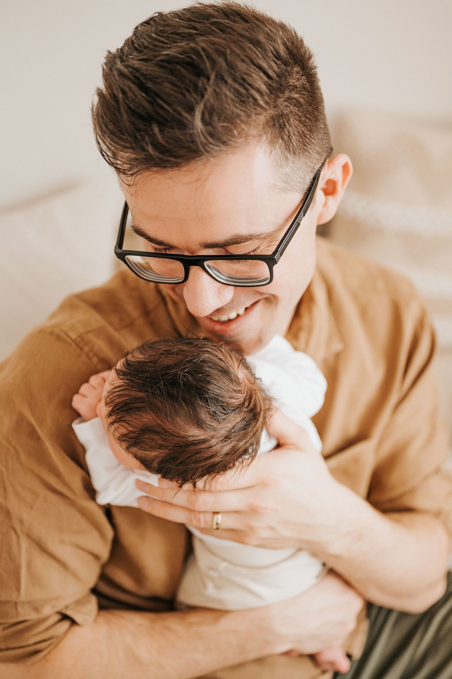 Parent wearing glasses and tan shirt holding newborn baby with tender expression of love and joy.