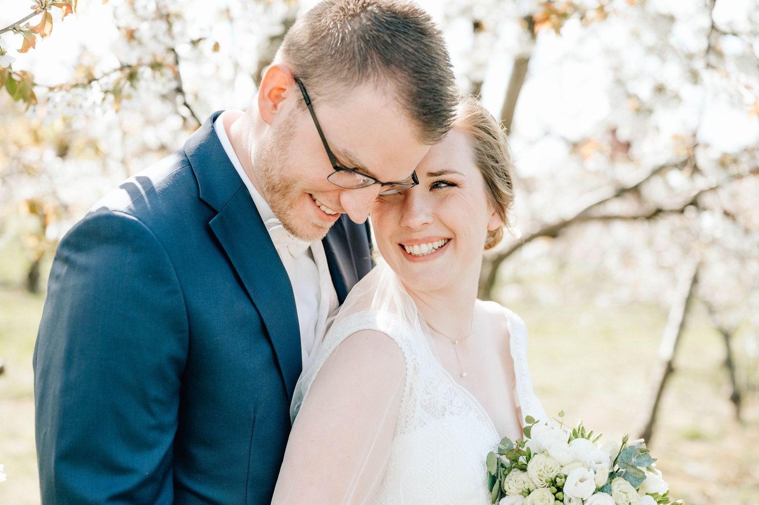 Joyful wedding portrait in an outdoor spring setting with white flowers and natural sunlight.