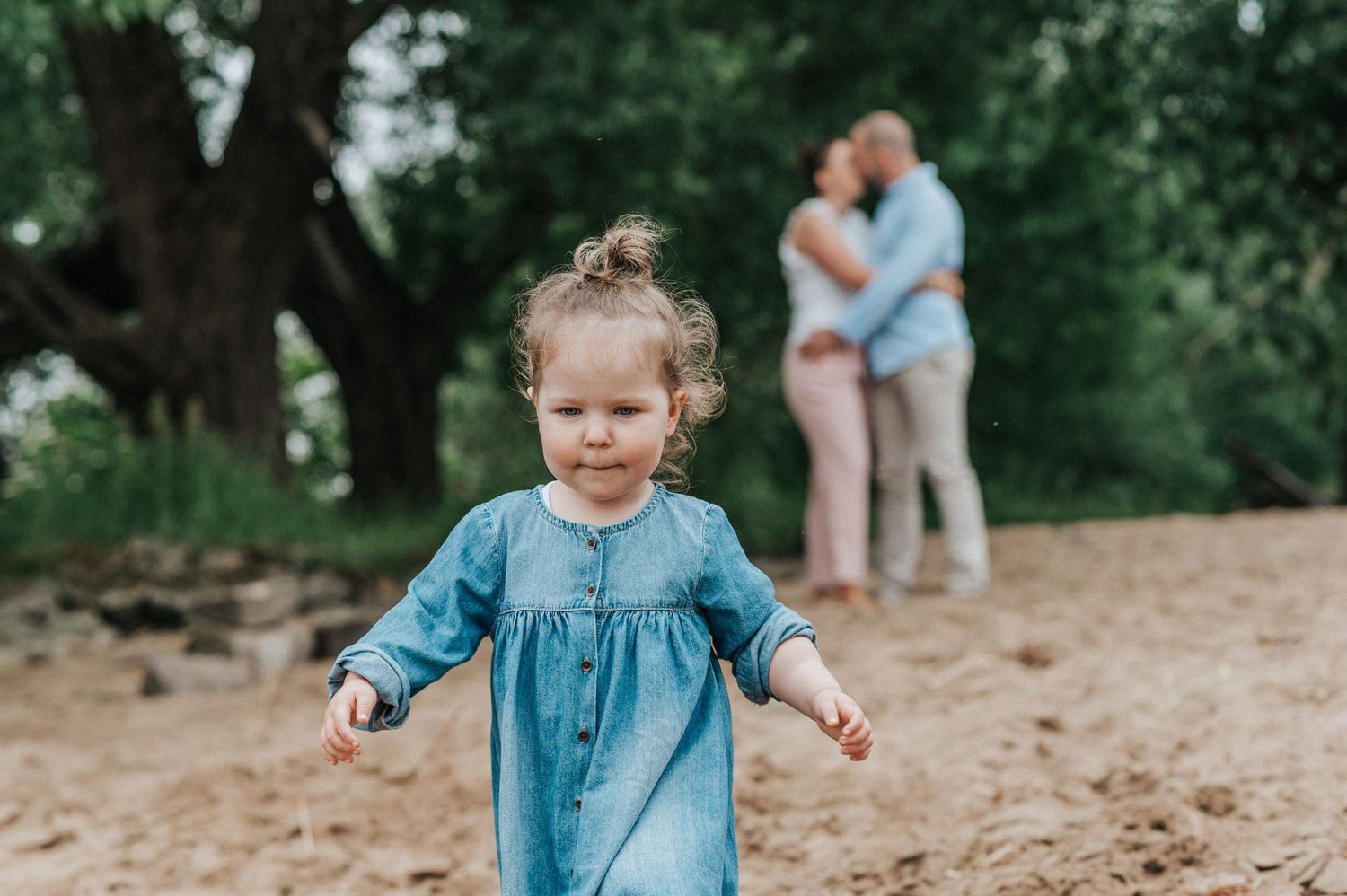 Child in blue dress playing outdoors with two people embracing in the background.