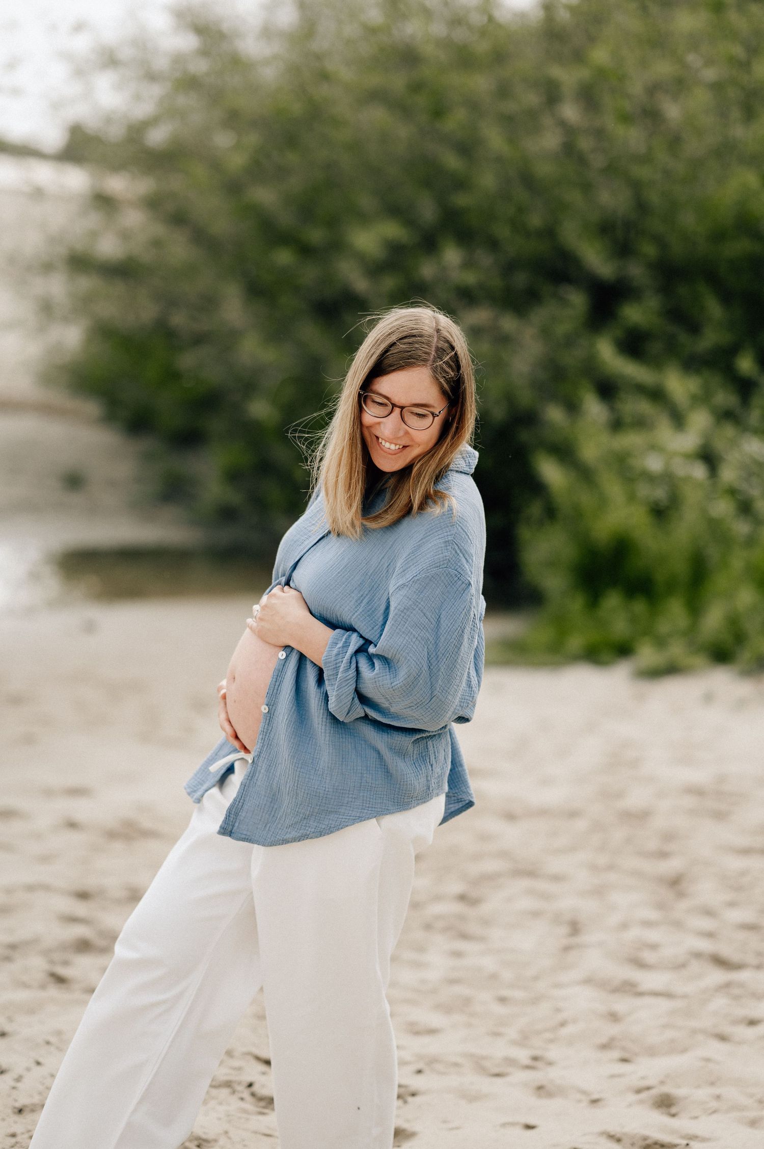 Expectant person in a casual blue sweater and white pants smiles while standing on a sandy beach.