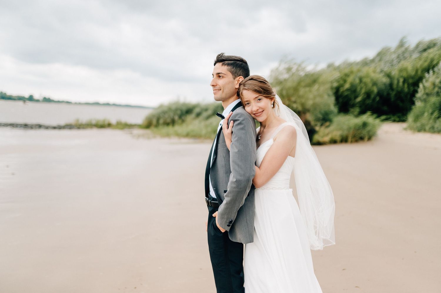 Newlywed couple in elegant attire standing together on sandy beach under cloudy sky.