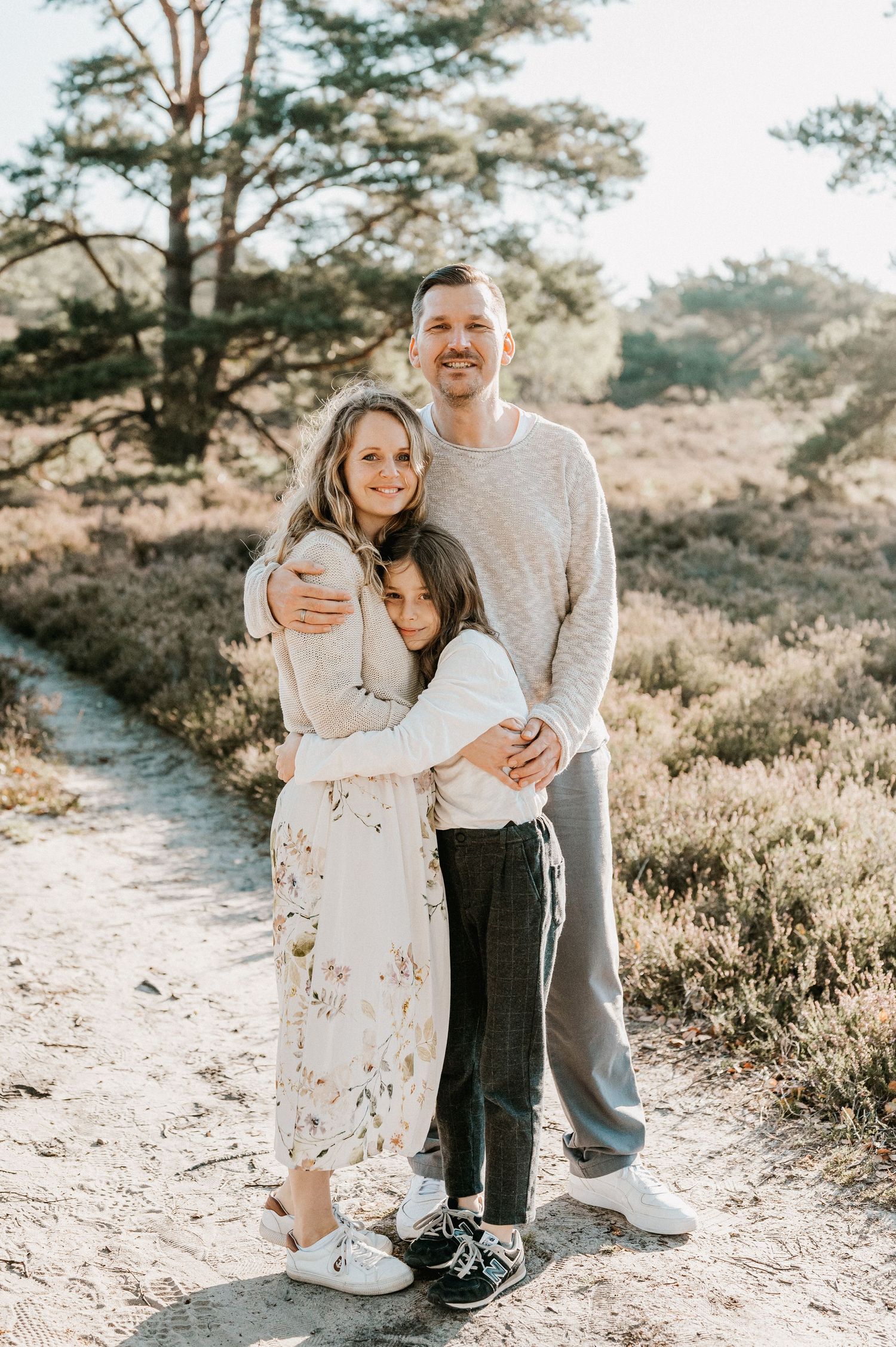 Family in light colored clothing embraces outdoors on a nature trail during golden hour sunset.
