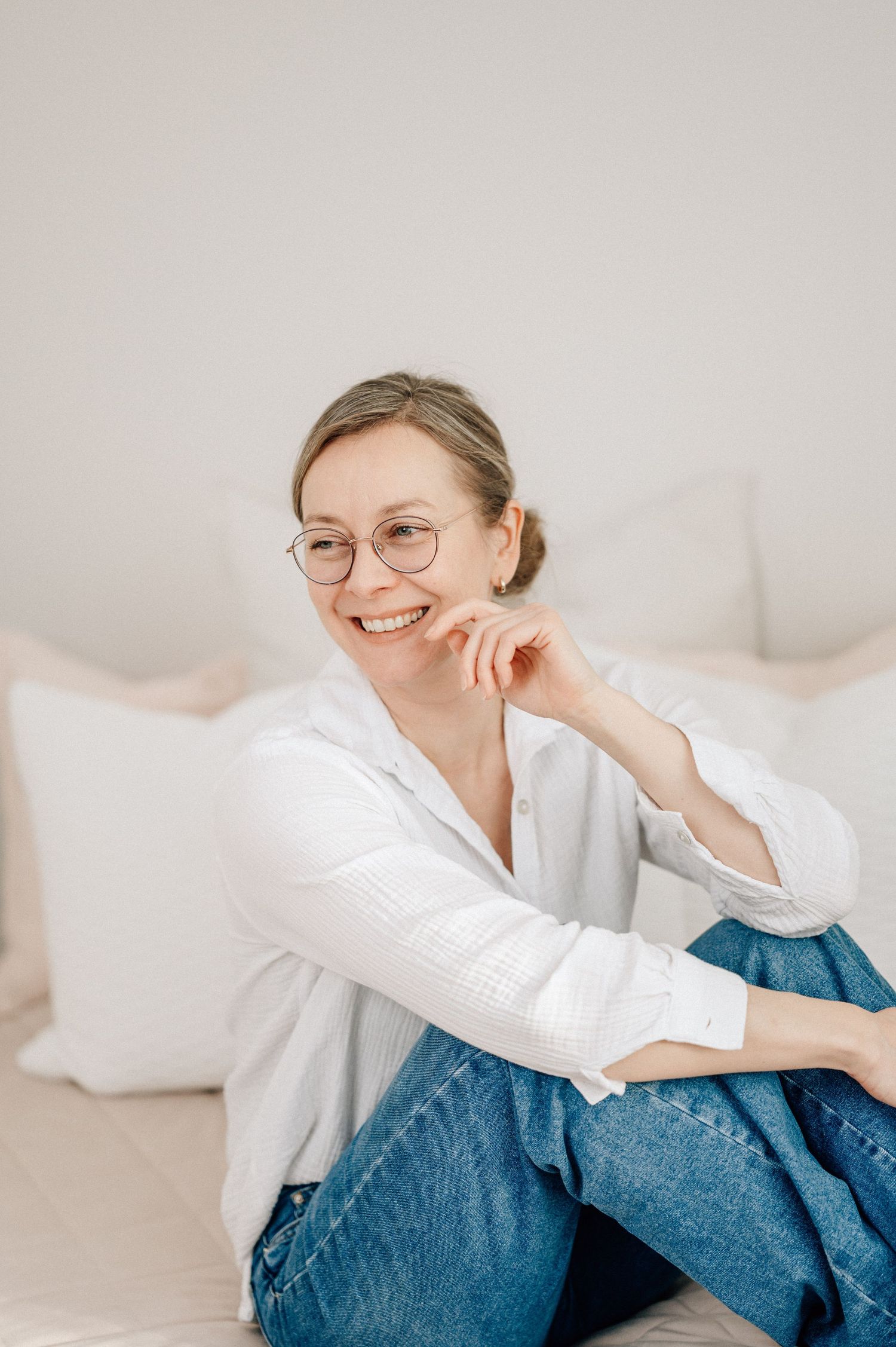 Smiling woman with glasses in white shirt and jeans sitting relaxed on a bed with white pillows.